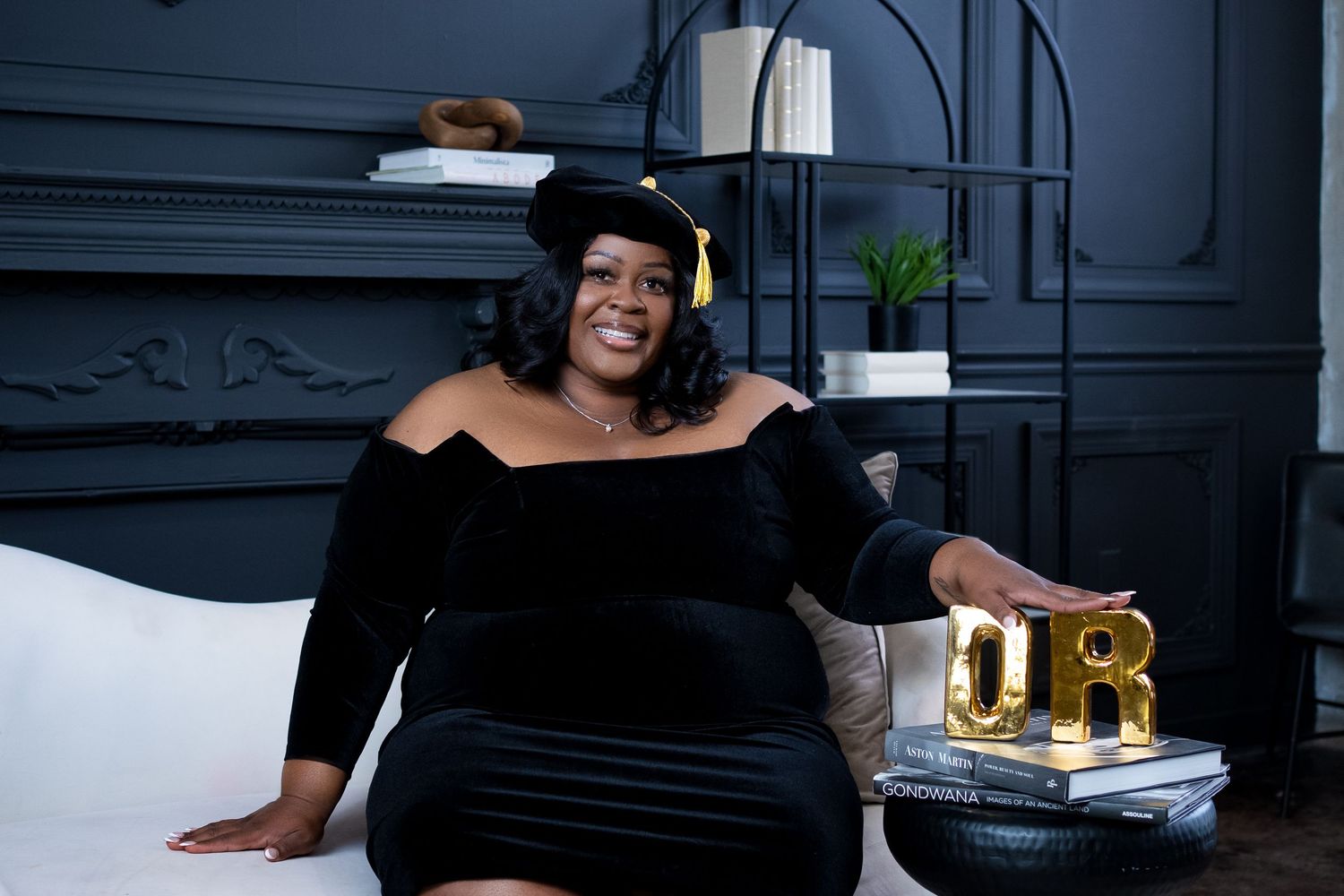 Graduate in elegant black gown poses on white couch with decorative 'DR' letters nearby.