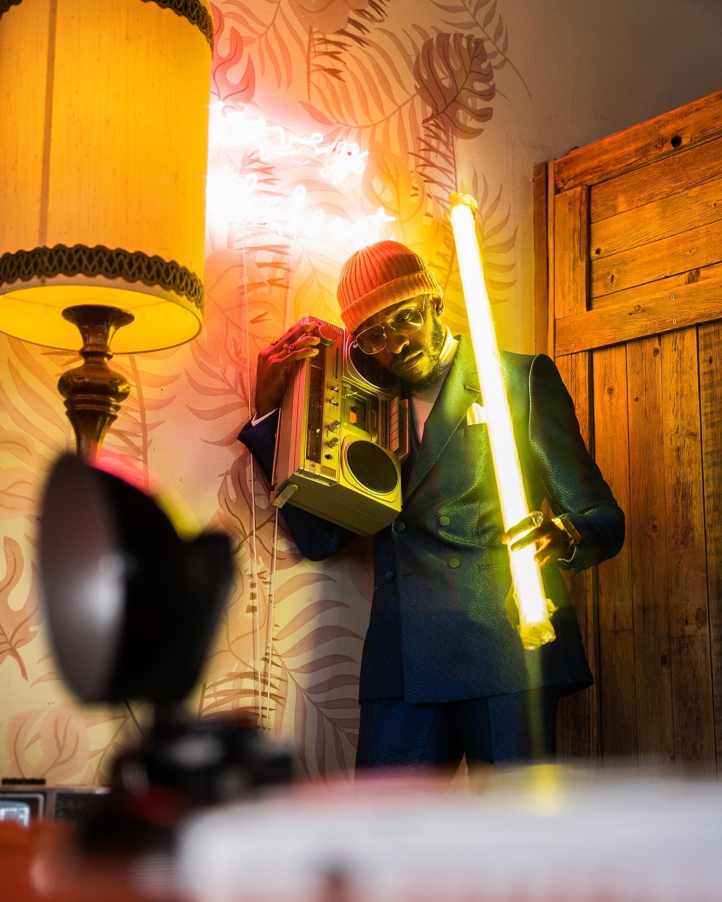 Person holding glowing neon lightsaber poses in room with vintage lamp and wallpaper.