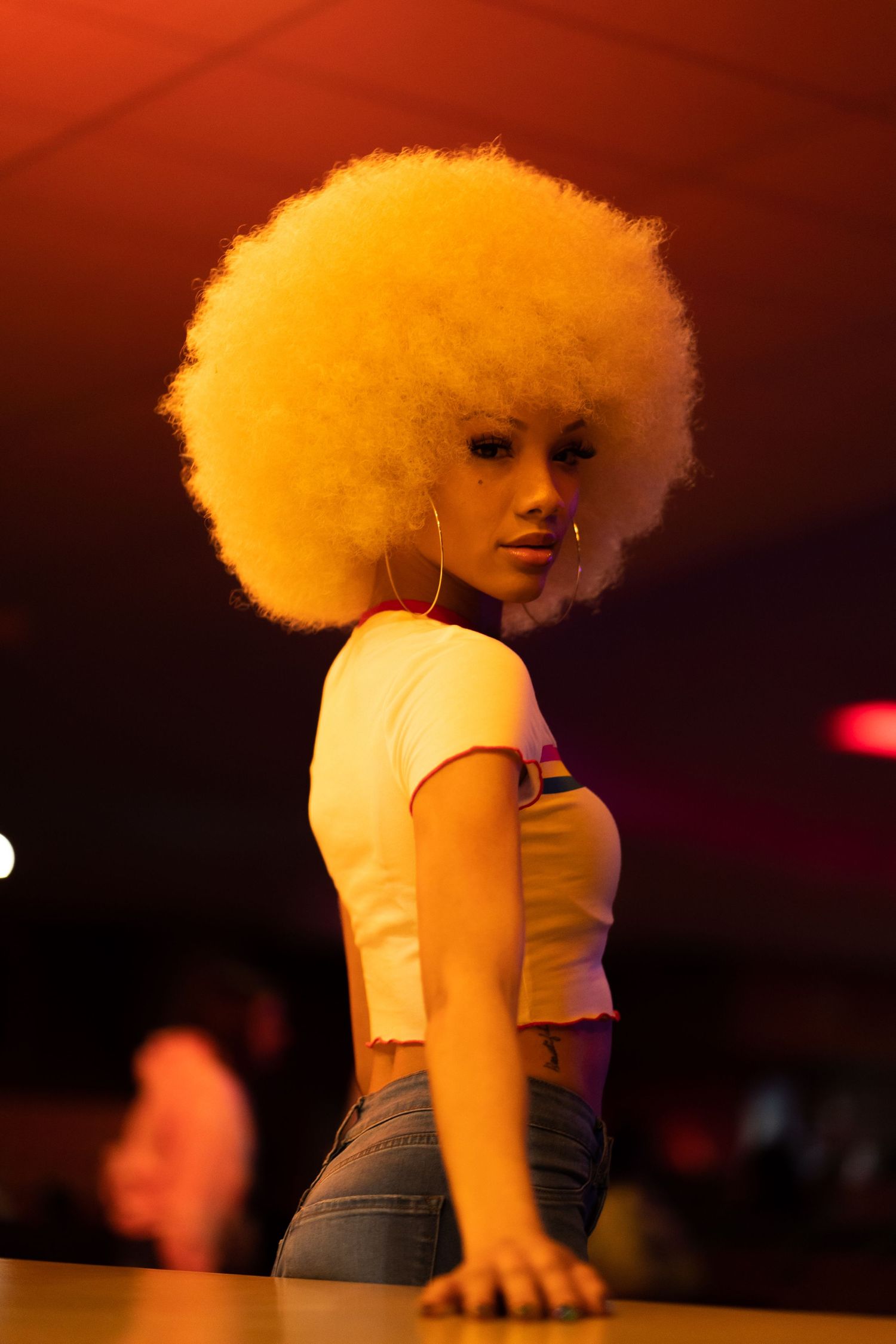 Model with large blonde afro poses on stage in retro-inspired outfit.