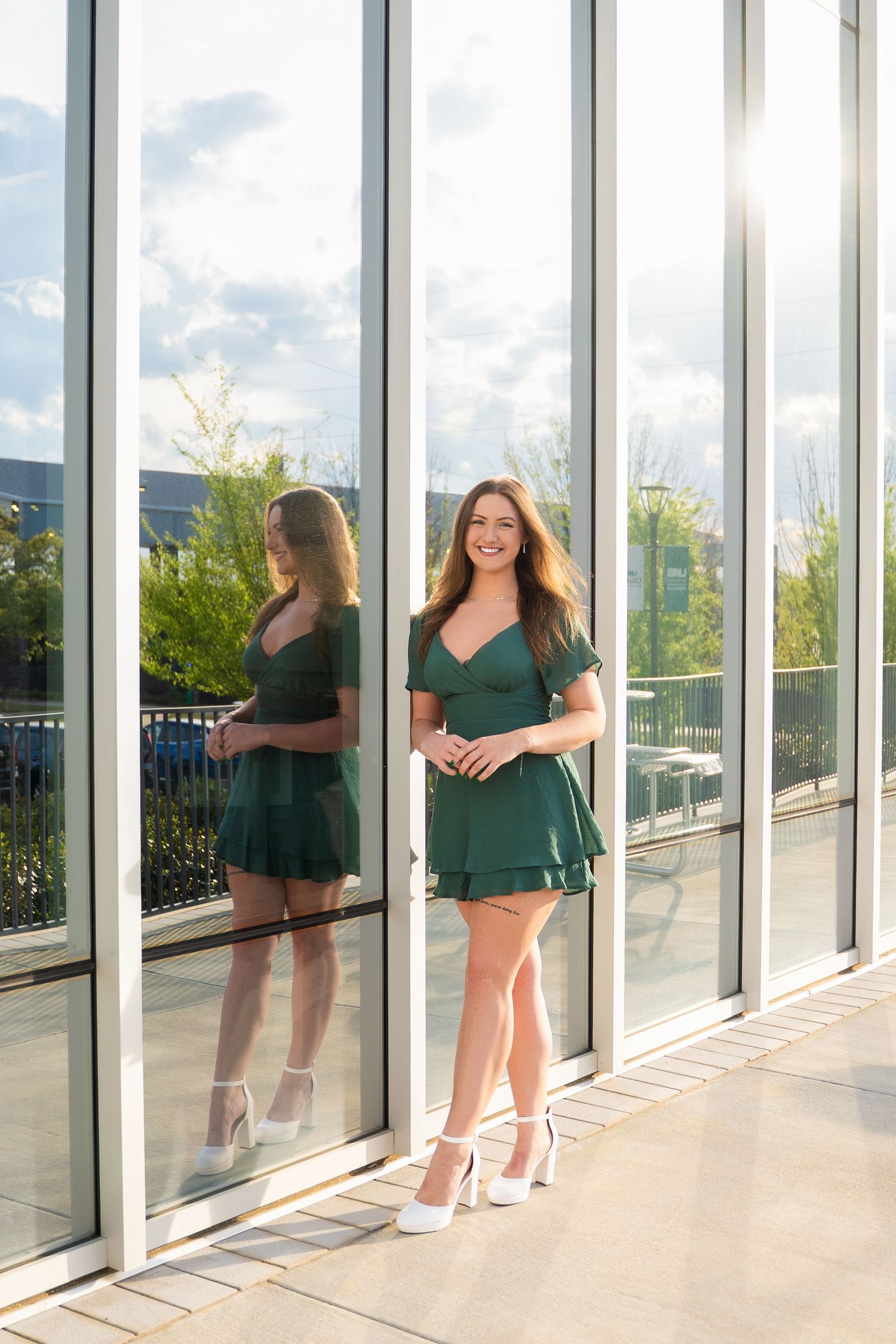Graduate in green dress poses against reflective glass windows in sunlight.