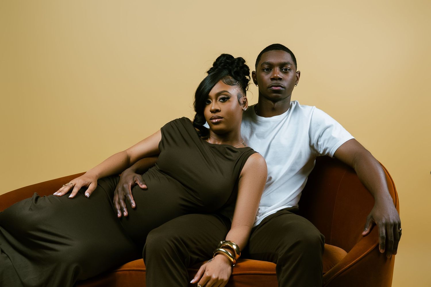 Maternity photo of couple relaxing on brown leather chair in casual attire.