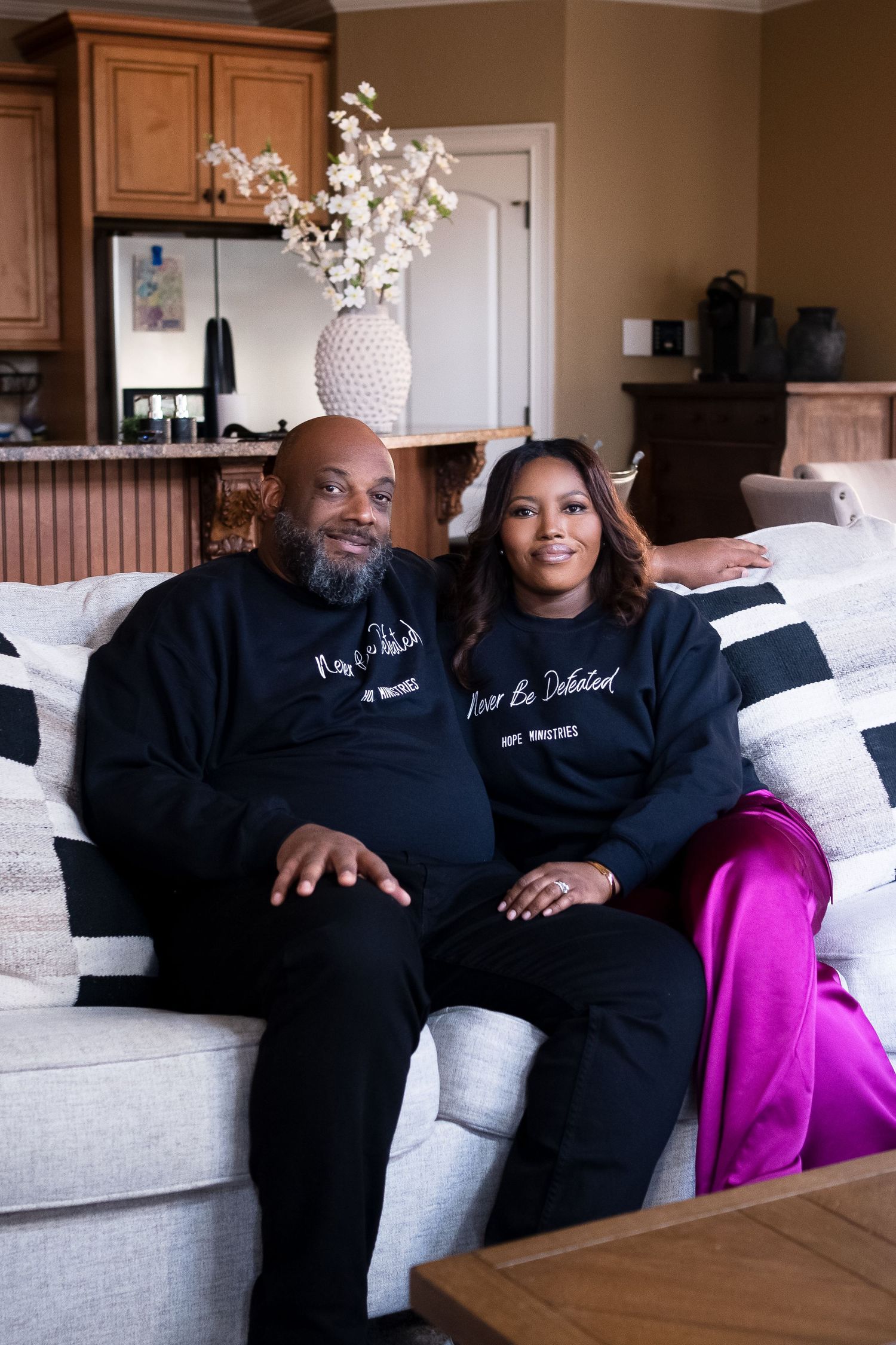 A couple wearing matching black sweatshirts relax together on a light-colored couch.