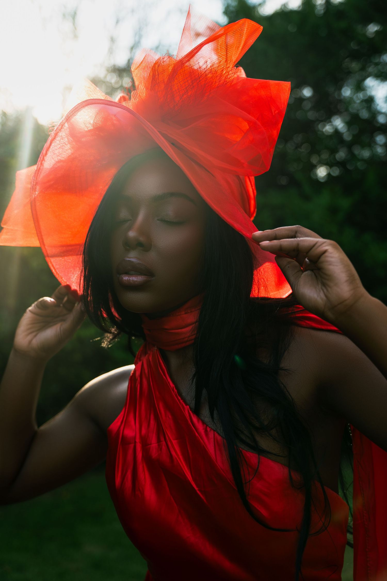 A figure in a flowing red dress and headpiece poses gracefully against a sunlit natural backdrop.