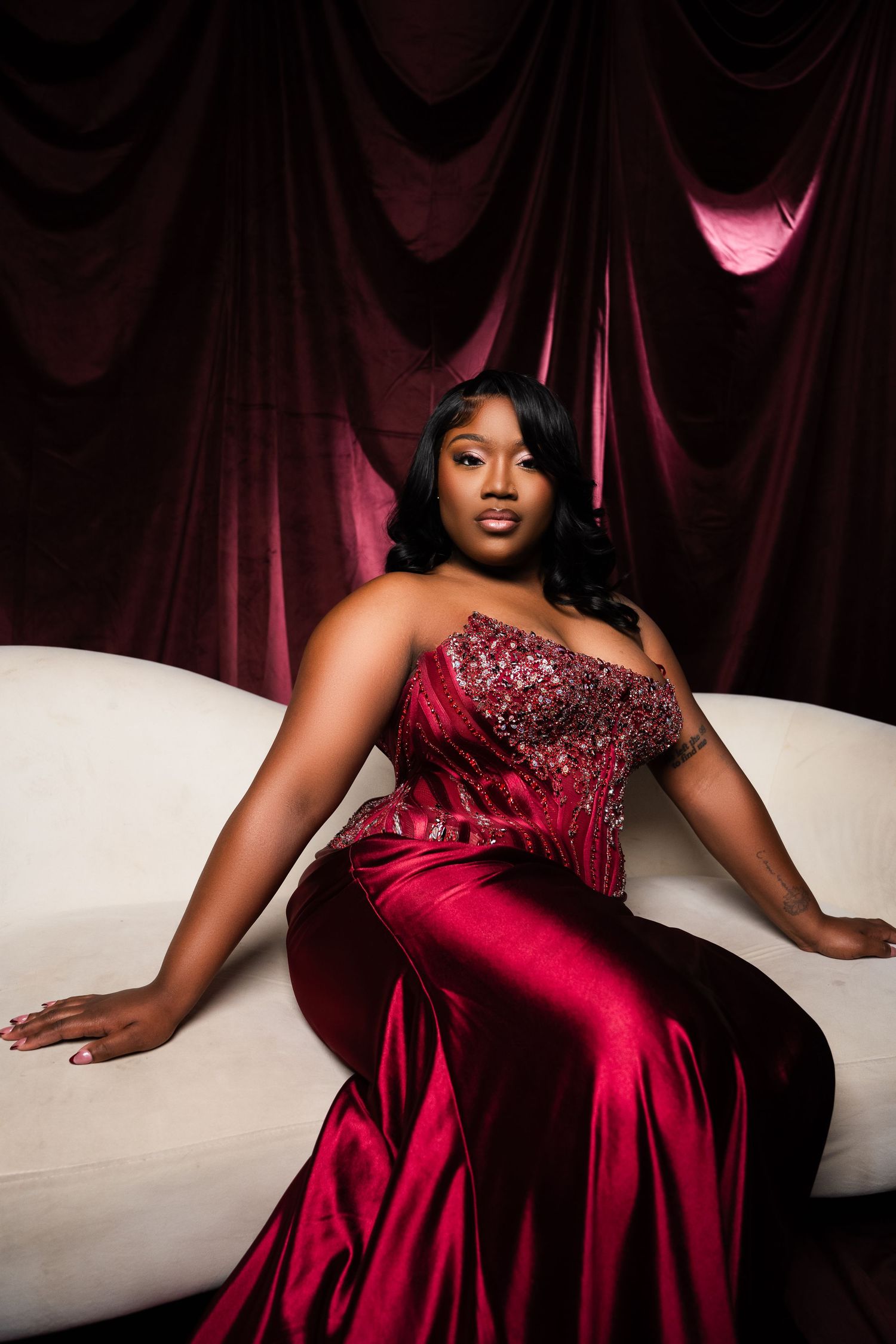 Woman in elegant red sequin strapless gown poses on white sofa against rich burgundy velvet drape backdrop.