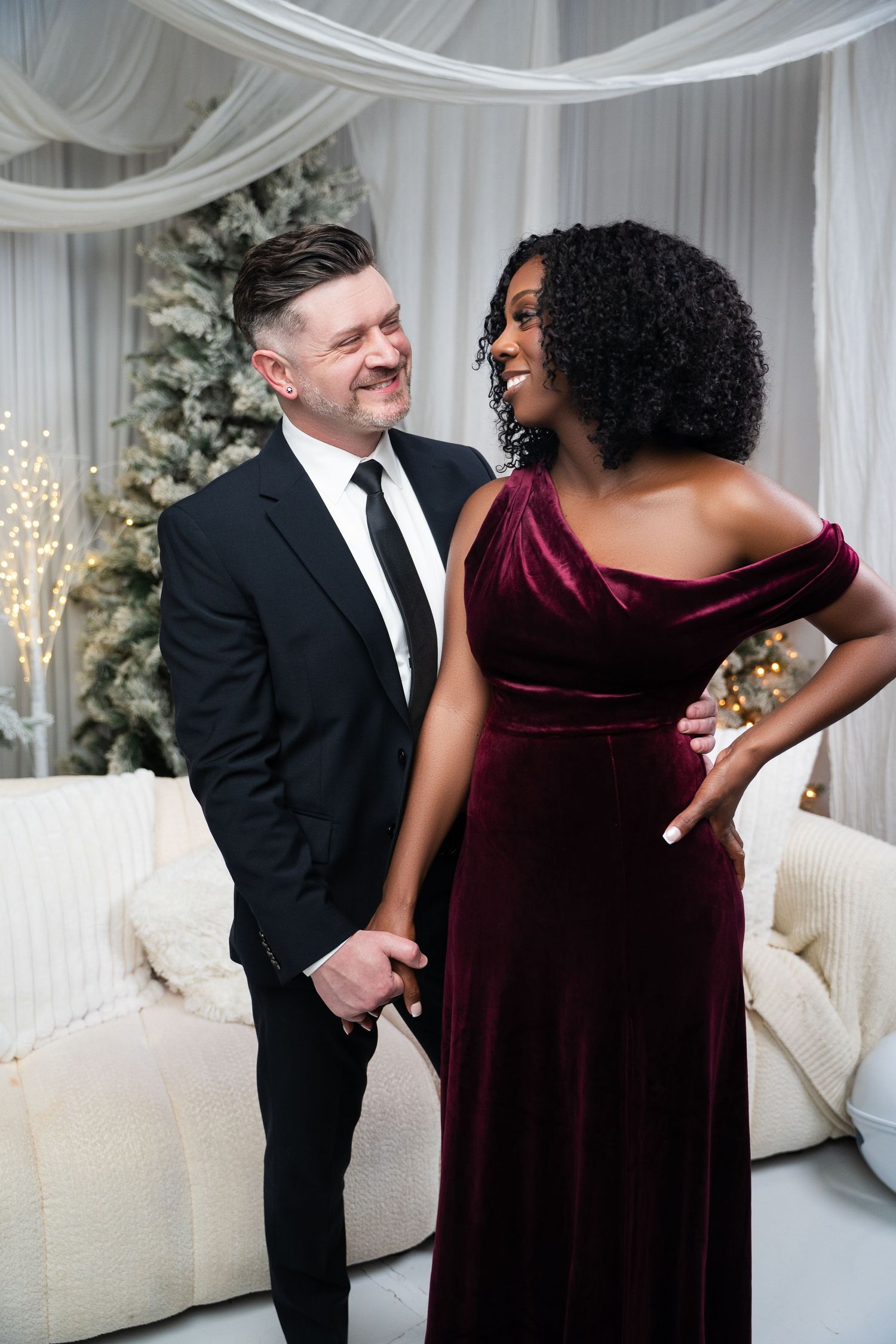 Interracial couple in elegant formalwear smiling at each other near a Christmas tree, woman in burgundy velvet gown, man in black suit.