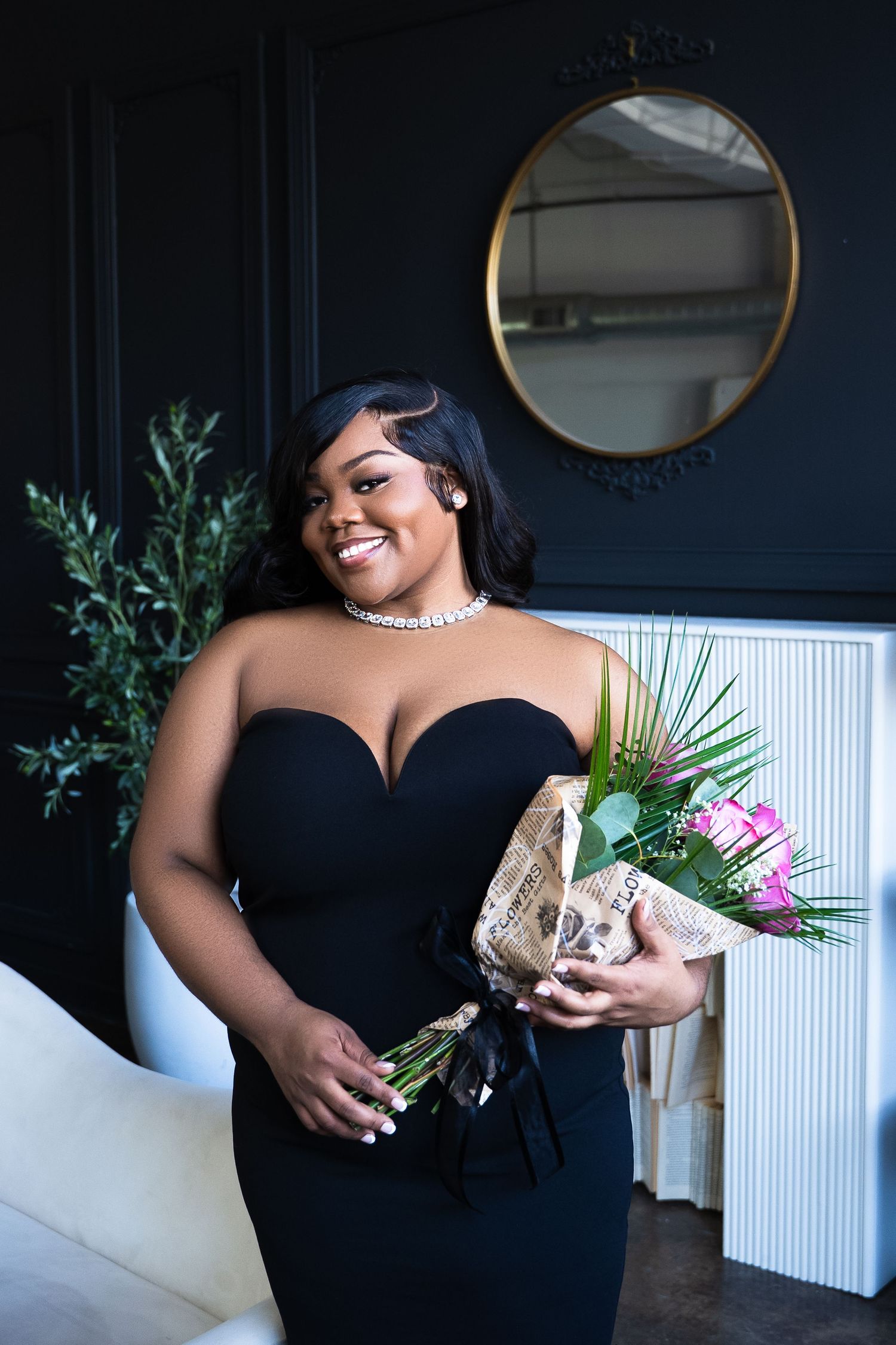 Elegant glamour portrait in black dress with flowers against dark wall with round mirror.