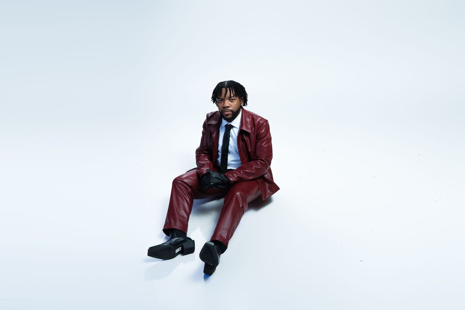 A seated figure wearing a burgundy suit against a white backdrop.