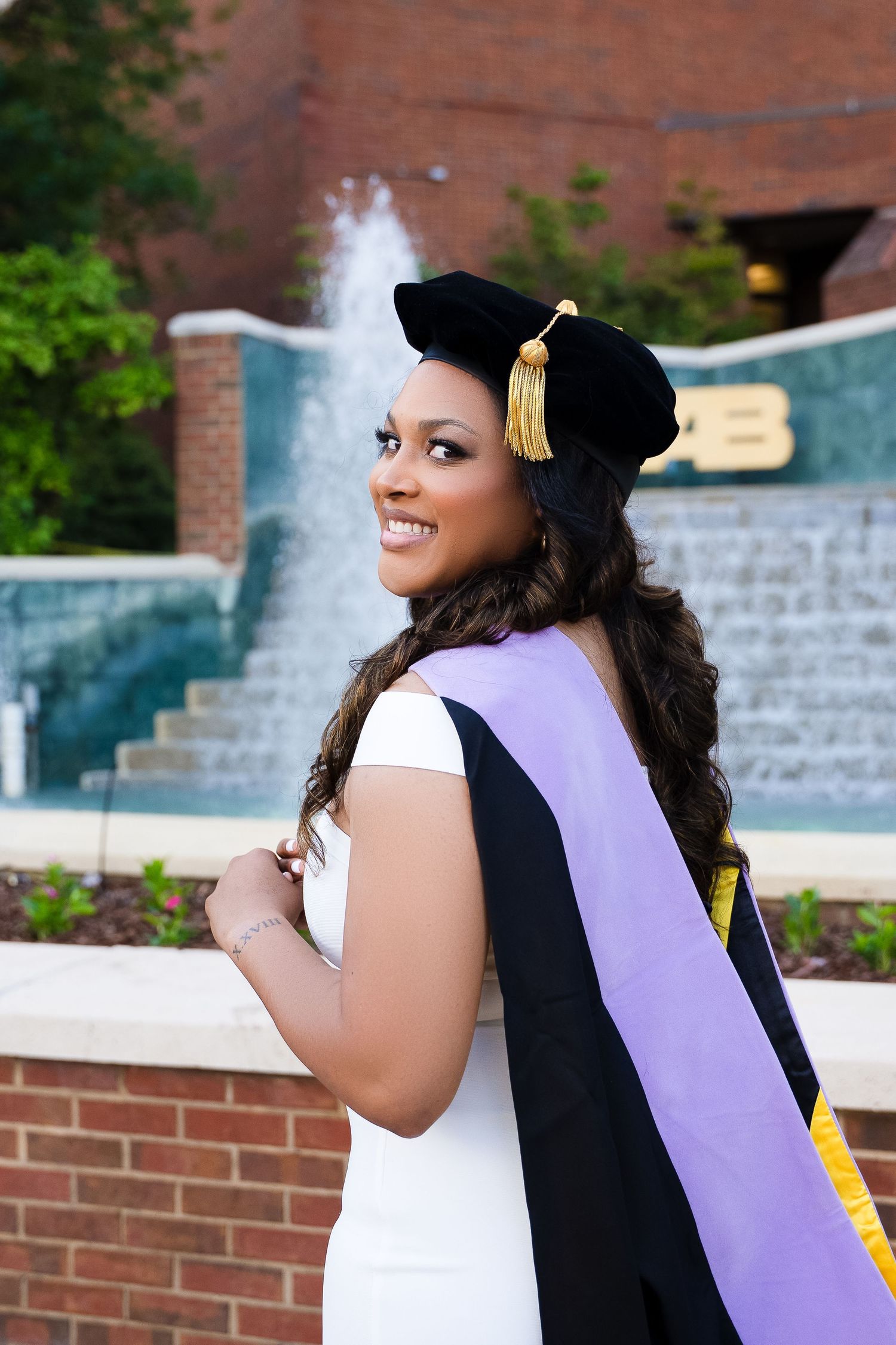 Graduate in cap and gown poses by campus fountain wearing purple graduation stole.