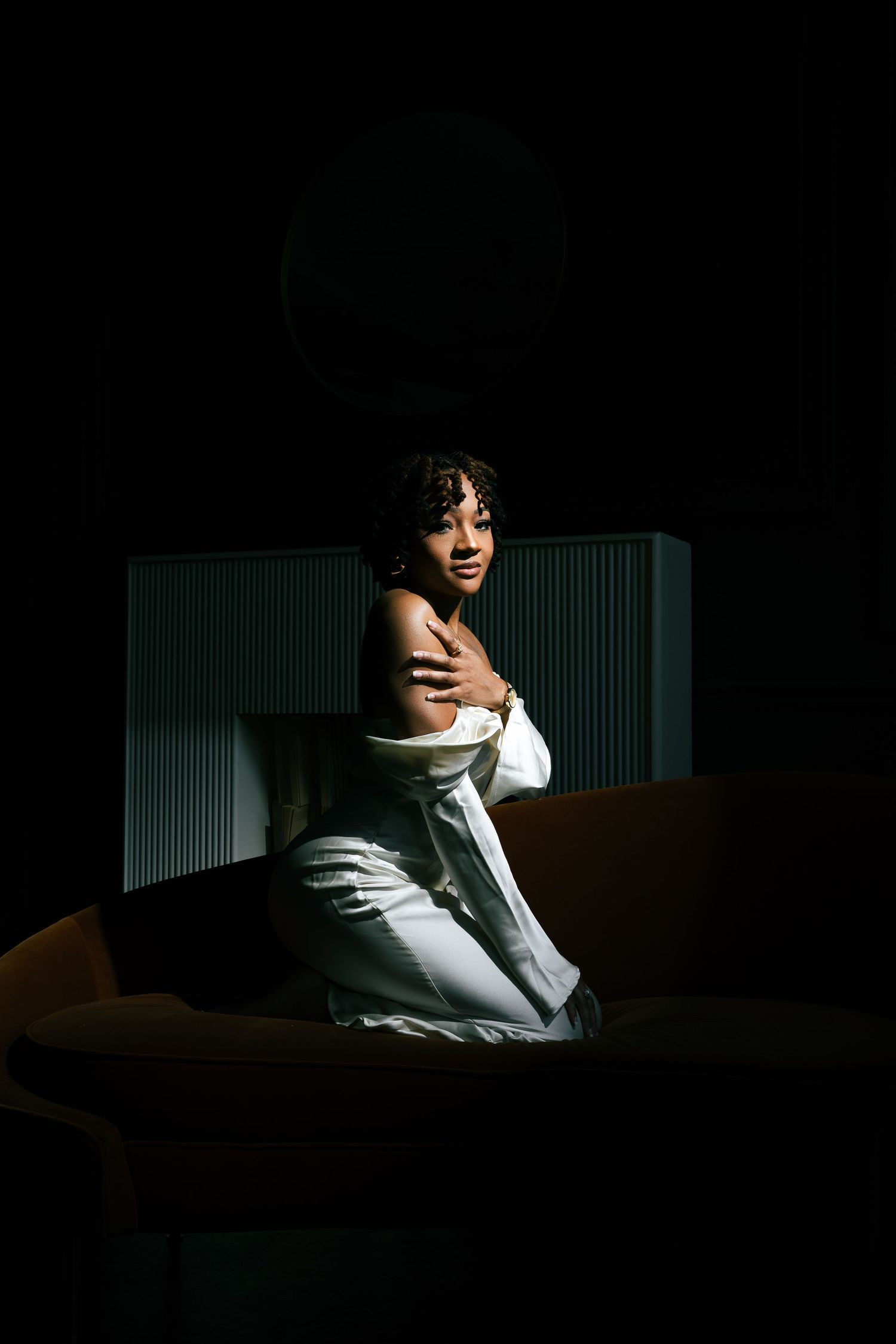A woman in a white shirt sits in a dimly lit room, hugging herself with a contemplative expression.
