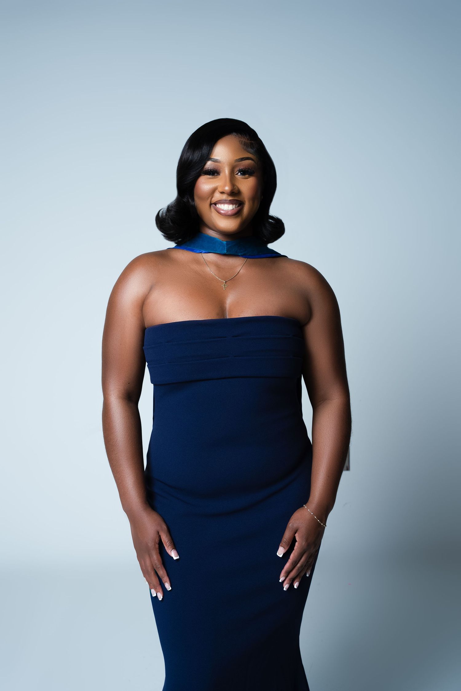 Woman in navy strapless gown with blue choker necklace smiling confidently against light gray background.