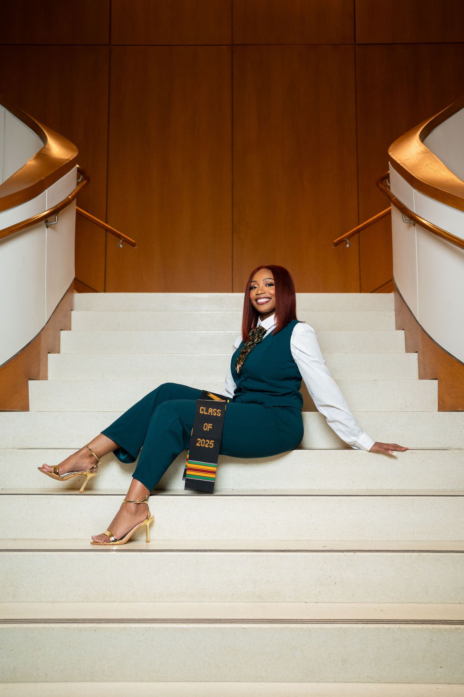 Professional portrait of someone sitting on white stairs wearing a dark green jumpsuit and white blouse with gold heels.