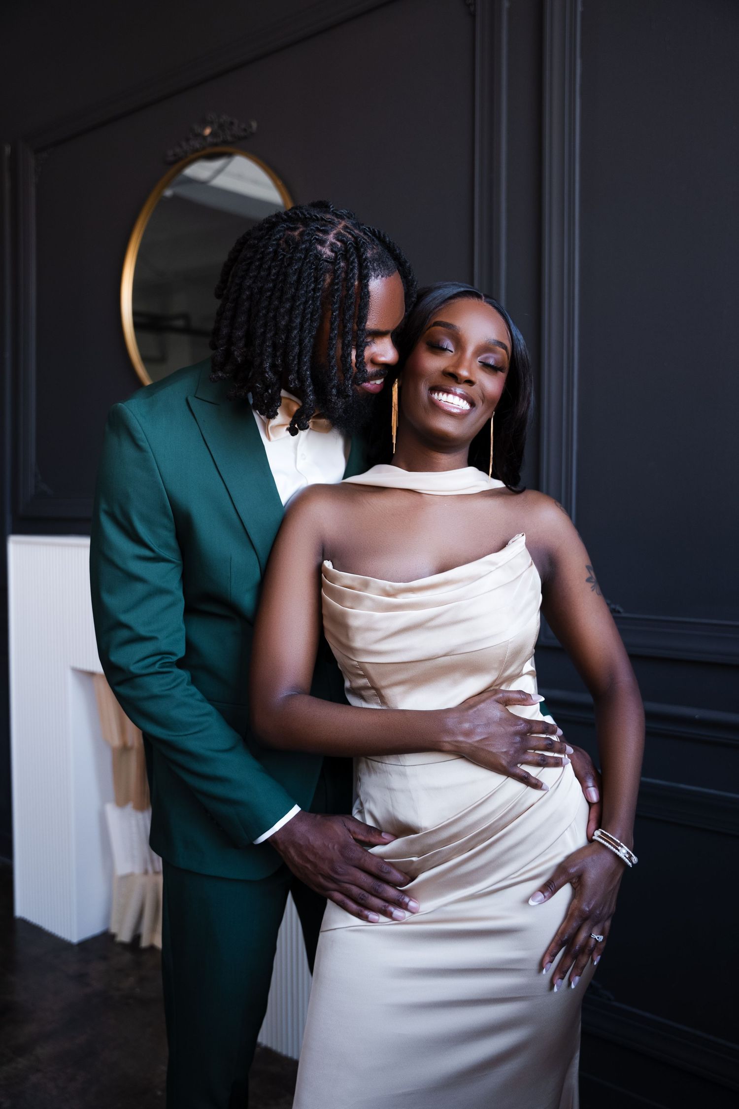Elegant couple posing together, man in green tuxedo embracing woman in champagne satin gown at formal event.