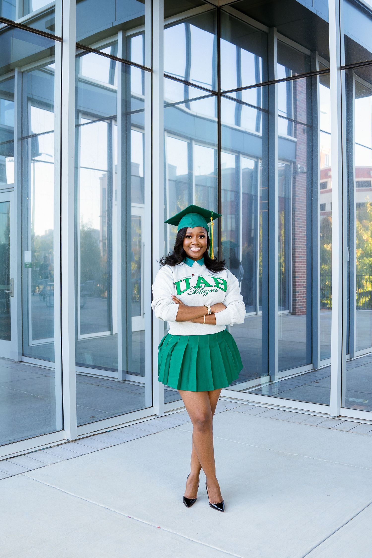 Graduate in green and white university attire poses in front of modern glass building.