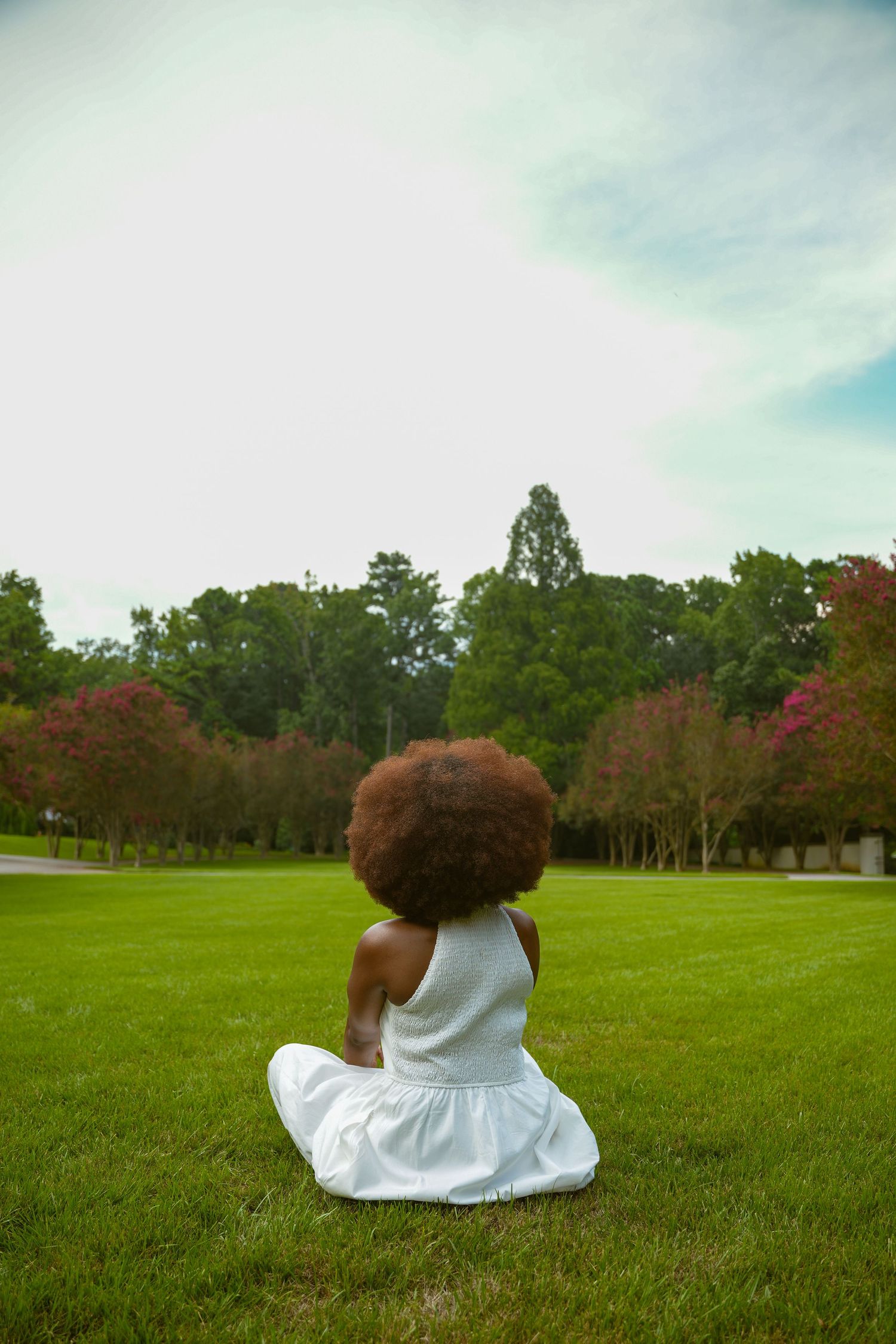 A figure in a white dress sits on a lush green lawn with trees and pink blossoms in the background.