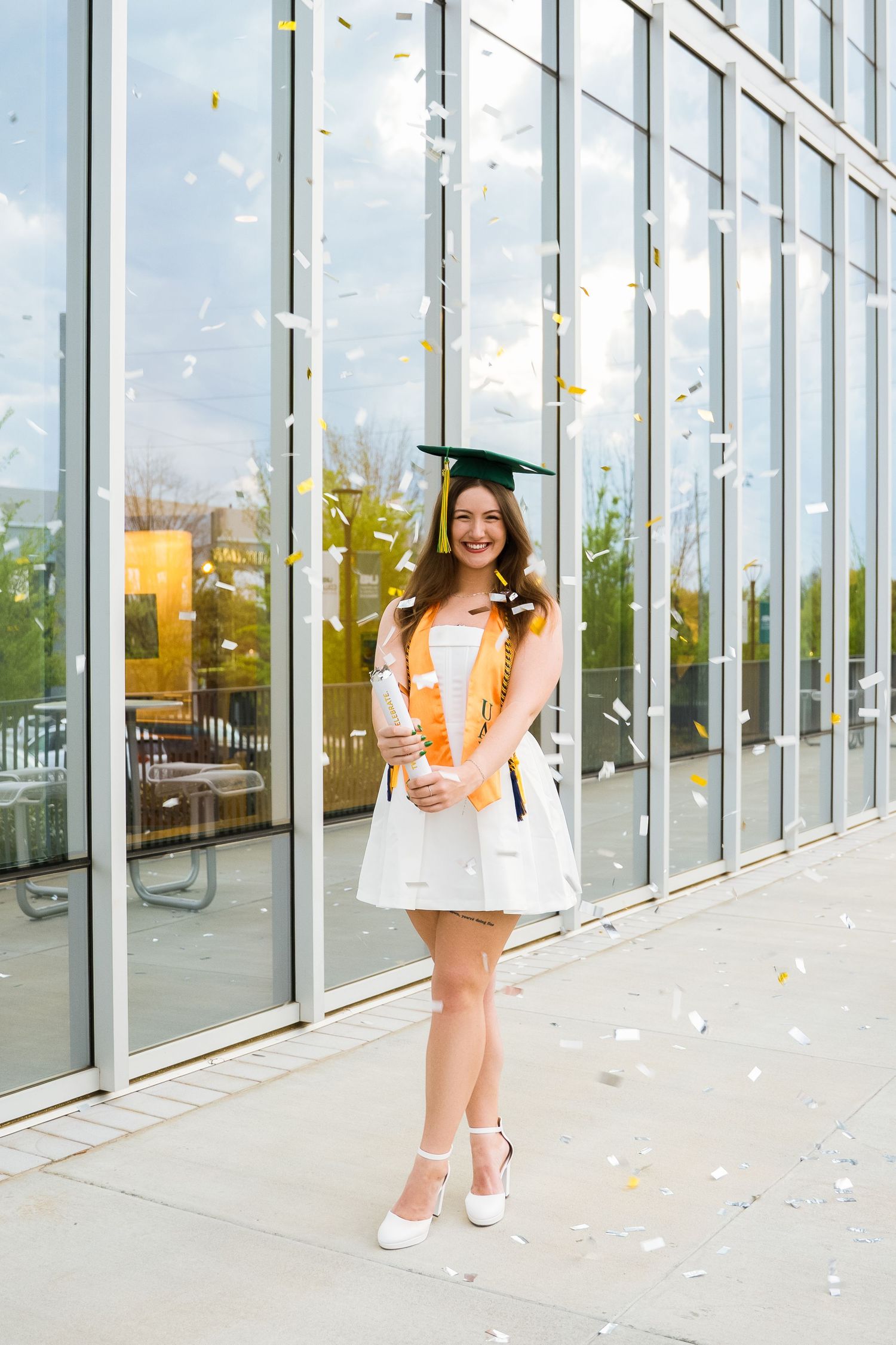 Graduate in white dress celebrates with confetti outside modern building.