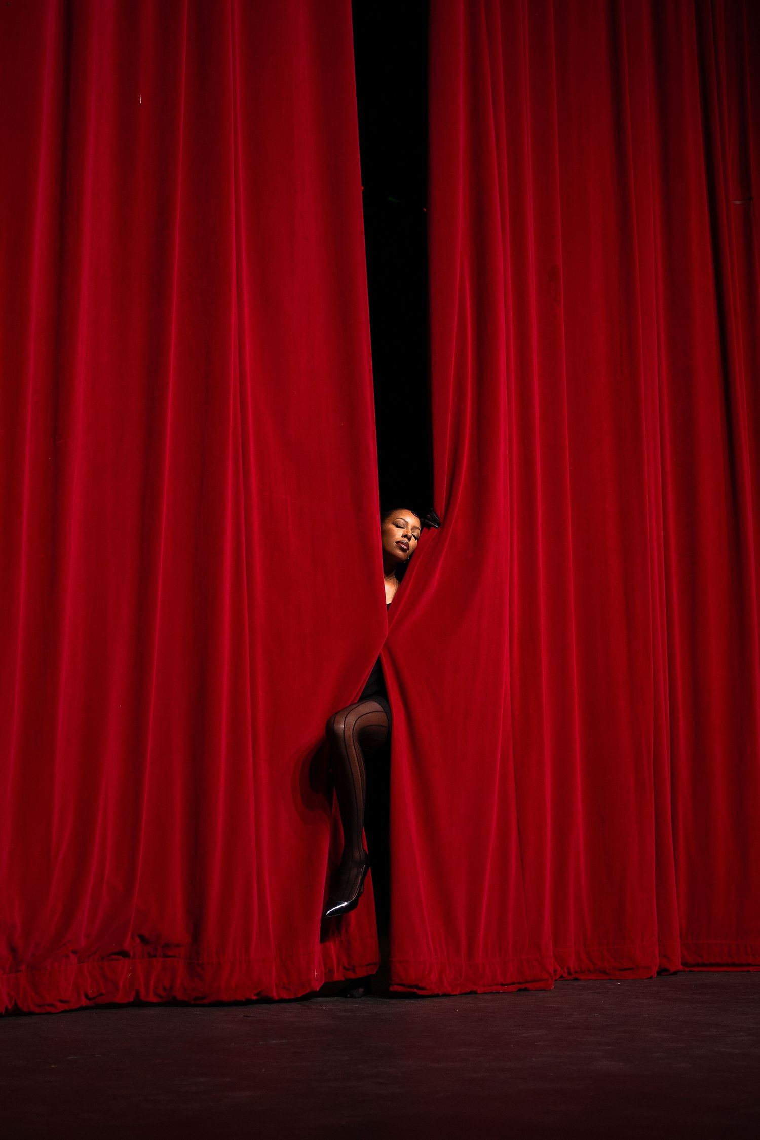 Dramatic figure peeks through red stage curtain in theatrical setting.