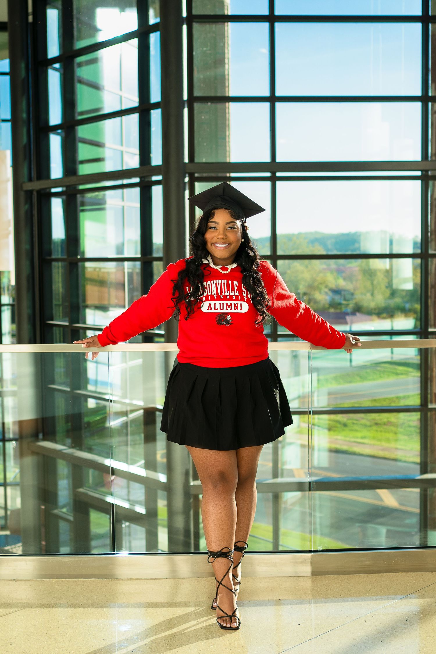 Graduate in red university sweatshirt and black skirt poses joyfully in modern building atrium.