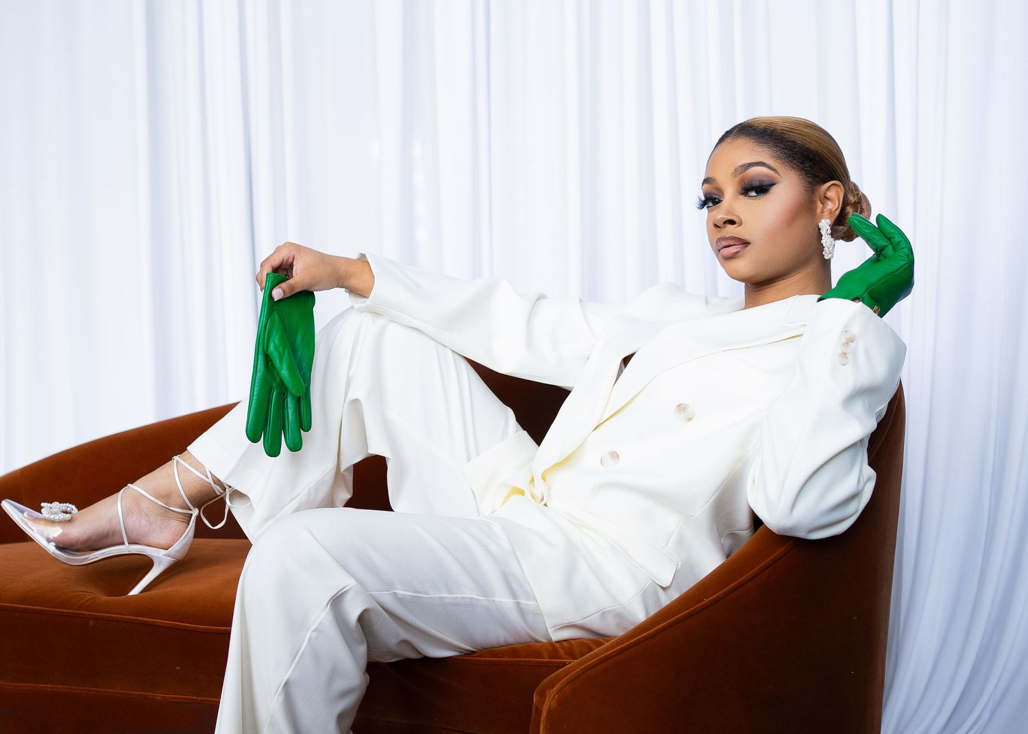 Modern fashion portrait in white suit with green gloves reclining on brown leather chair.