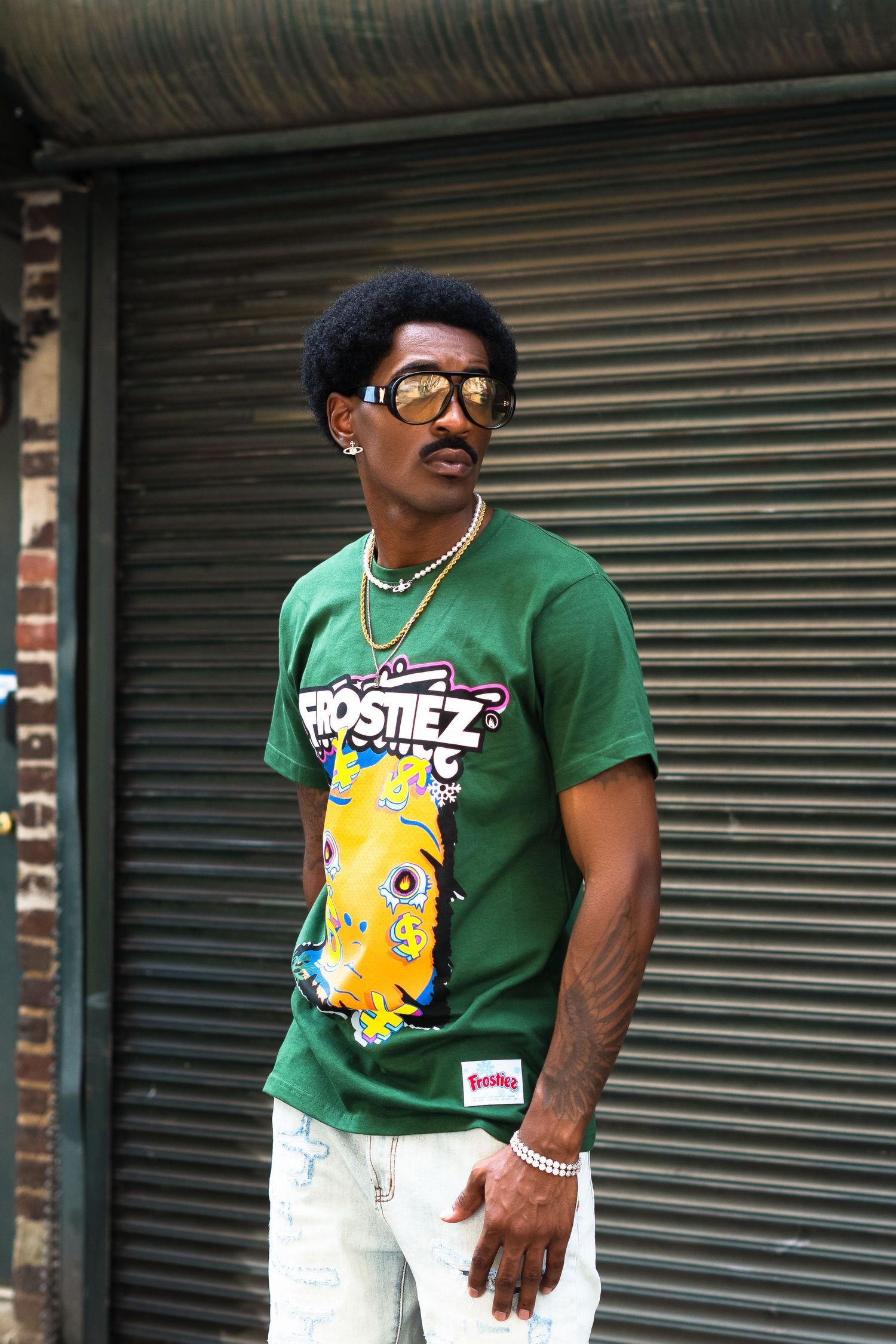 Stylish streetwear fashion shot of person in green graphic t-shirt with gold chain and sunglasses against metal door.