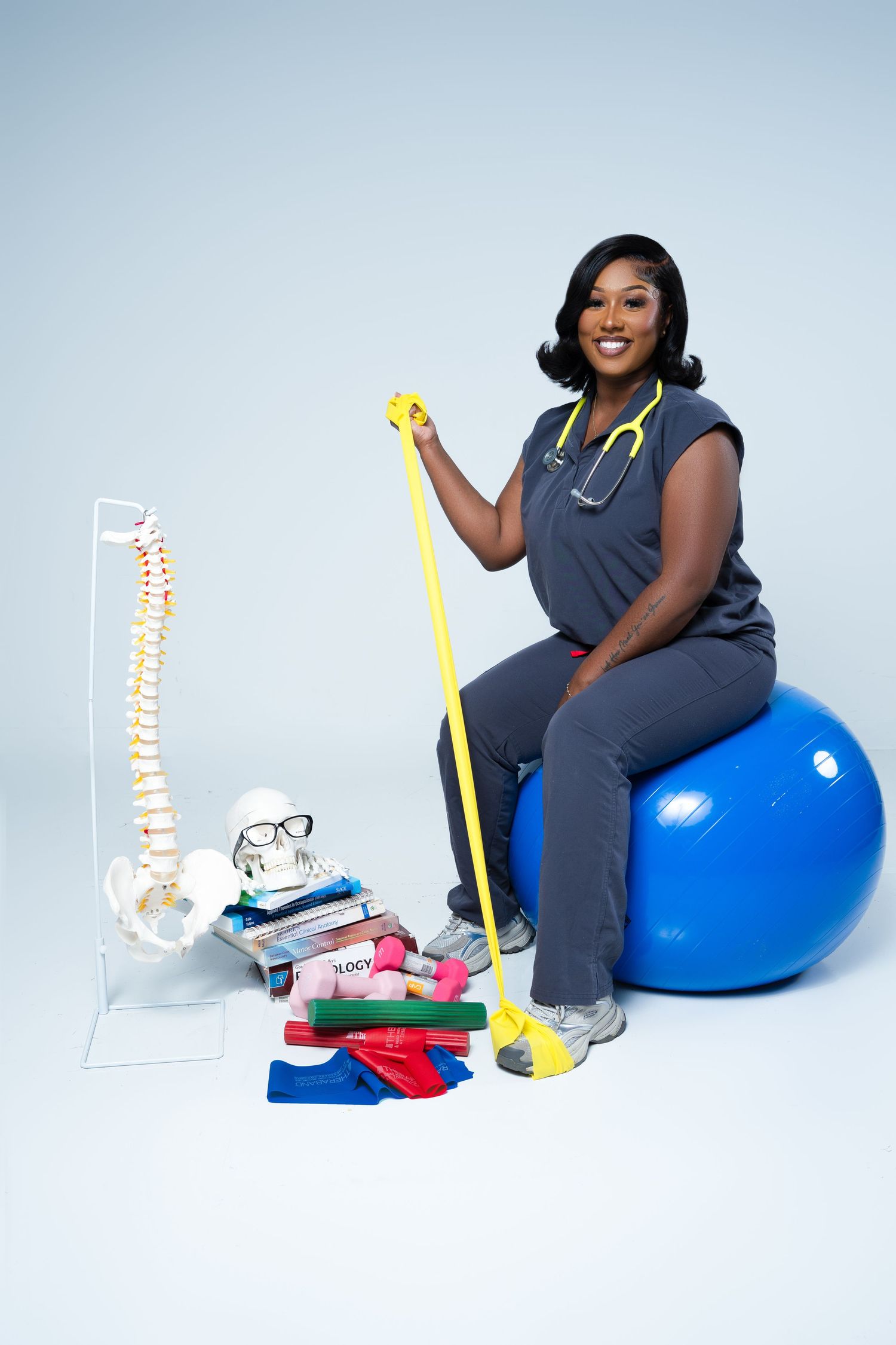 Female physical therapist in scrubs sits on blue exercise ball, holding resistance band near spine model and therapy equipment.