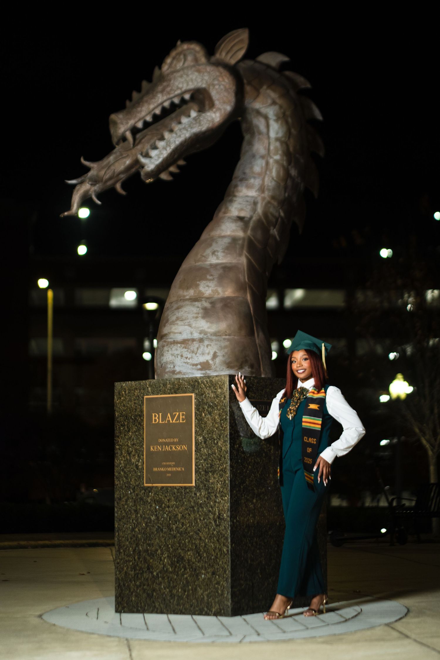 A skateboarder performs tricks near a large bronze dragon statue at night in a park.