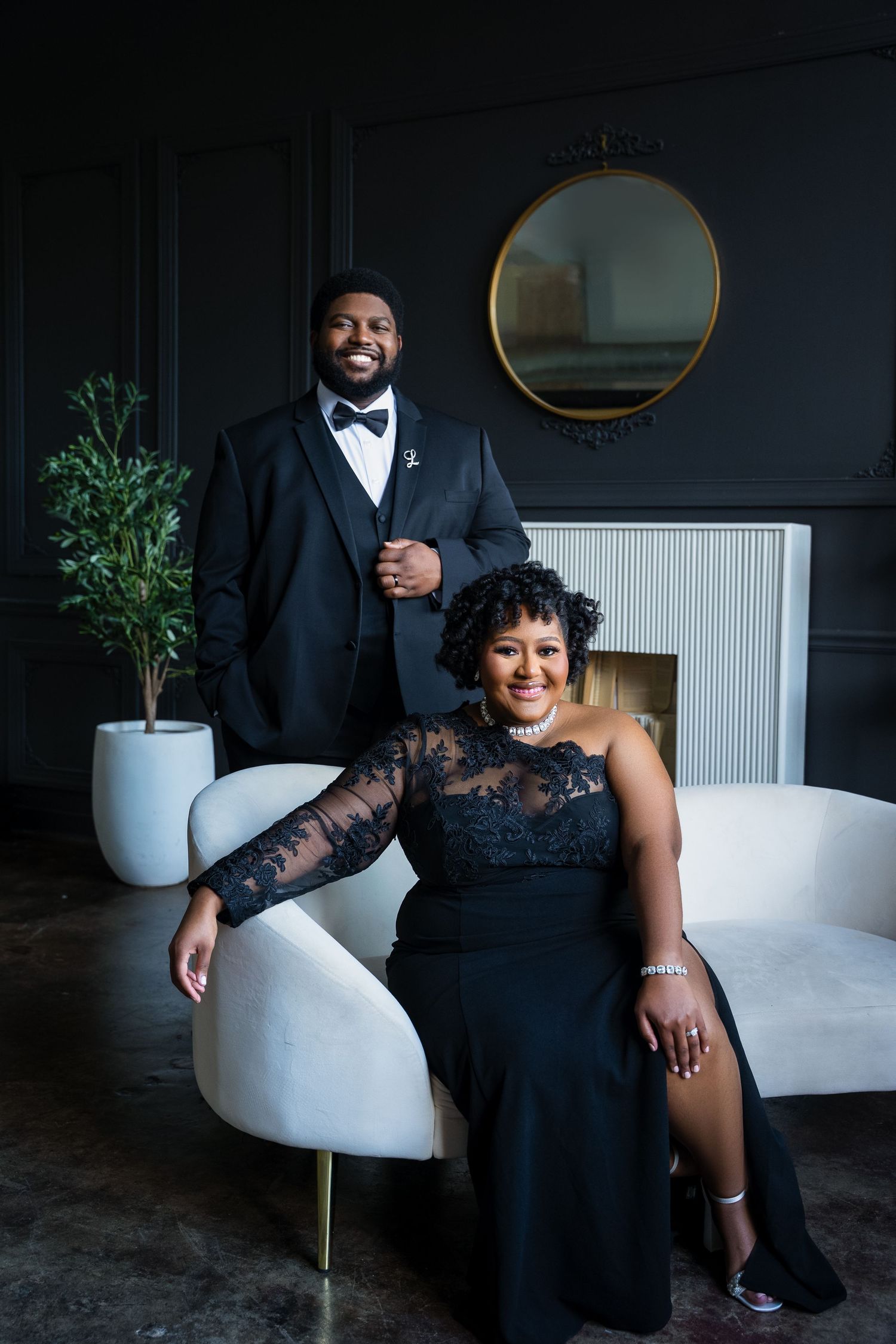 A couple in formal black attire poses together on a modern white chair against a dark backdrop with greenery.