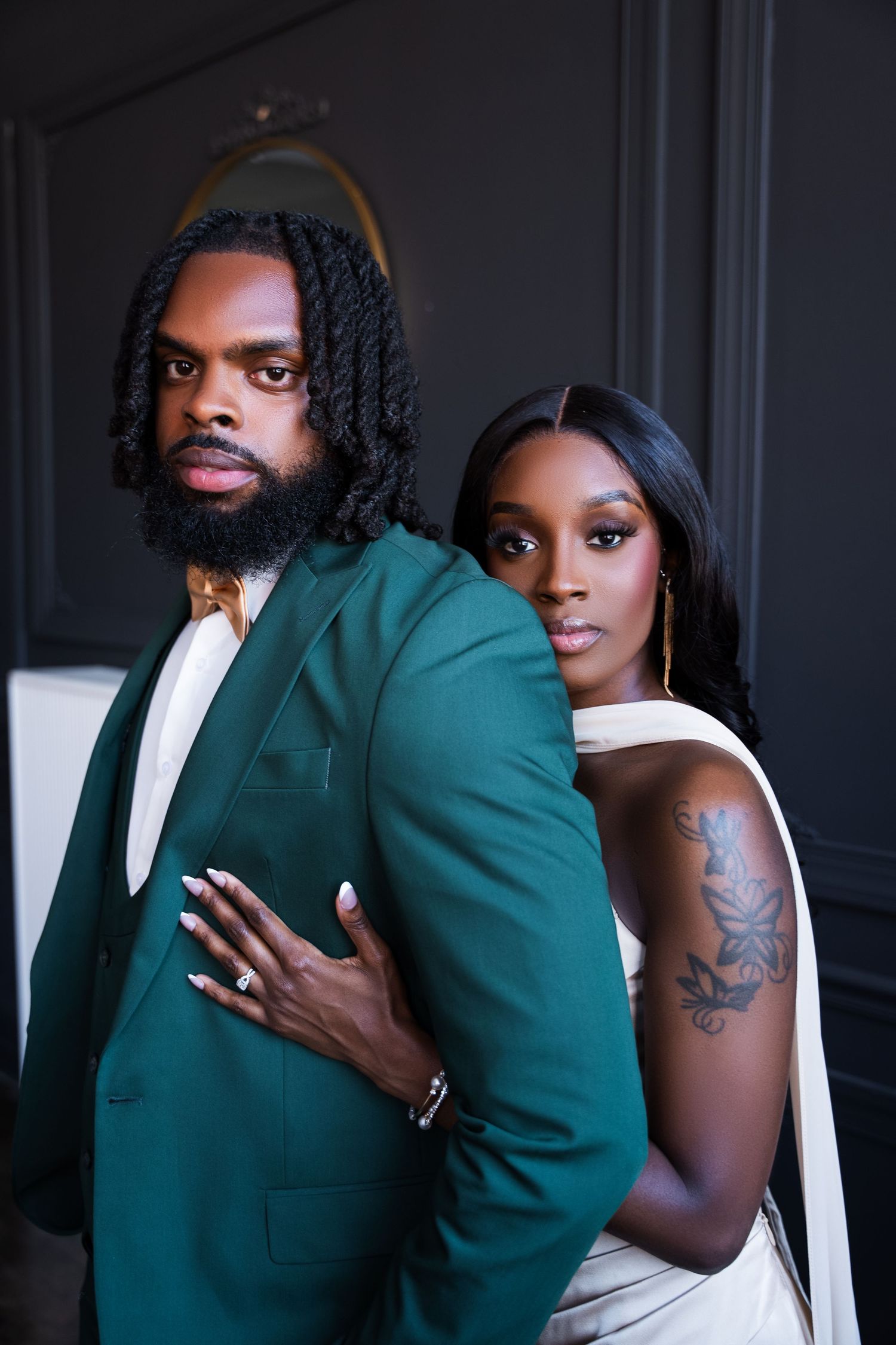 A stylish couple poses together, man in green suit with locs and beard, woman in white dress with floral tattoo.