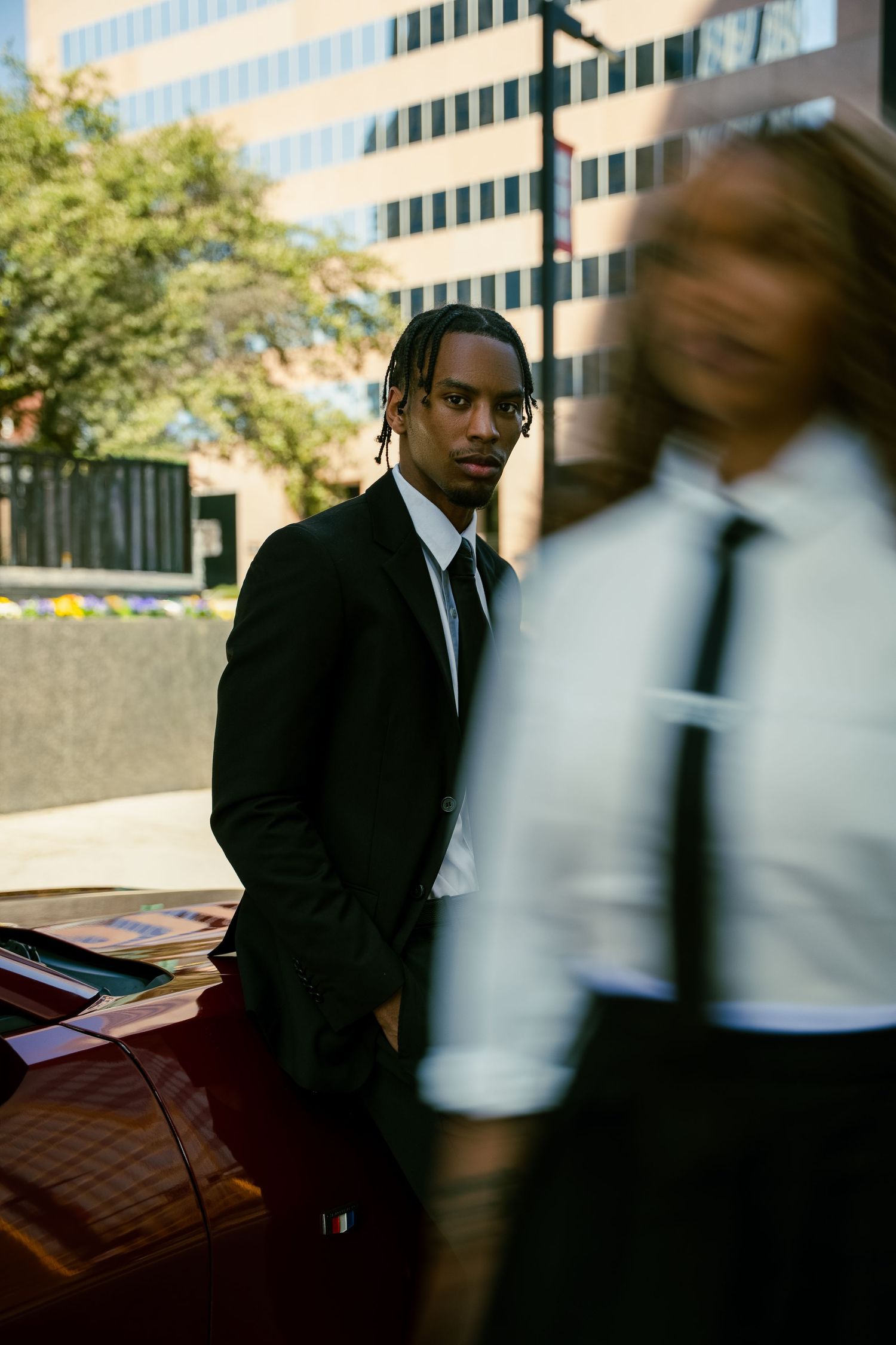 A person in a dark business suit stands near an office building while someone in white walks past in a blurred motion.
