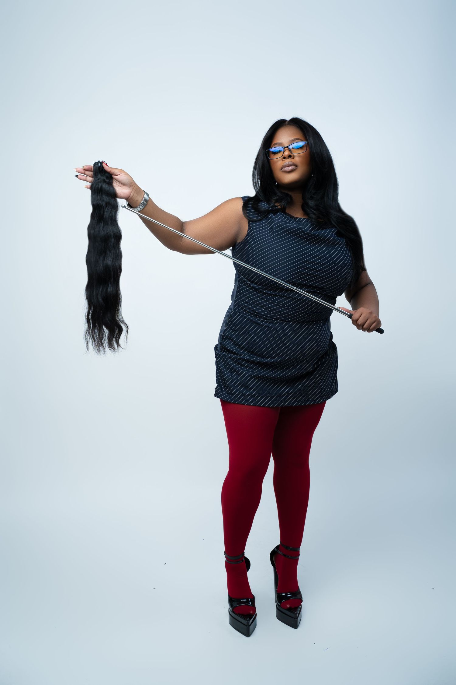 Woman in blue dress and red tights holds hair extensions and a pointer stick against white background.