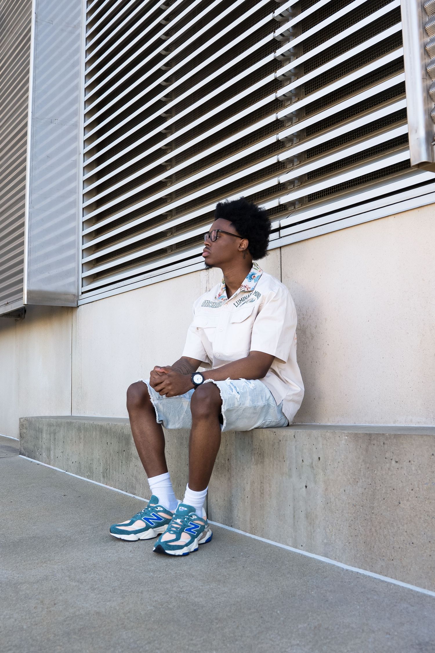 Urban fashion portrait with person in white shirt and blue sneakers sitting against industrial building.