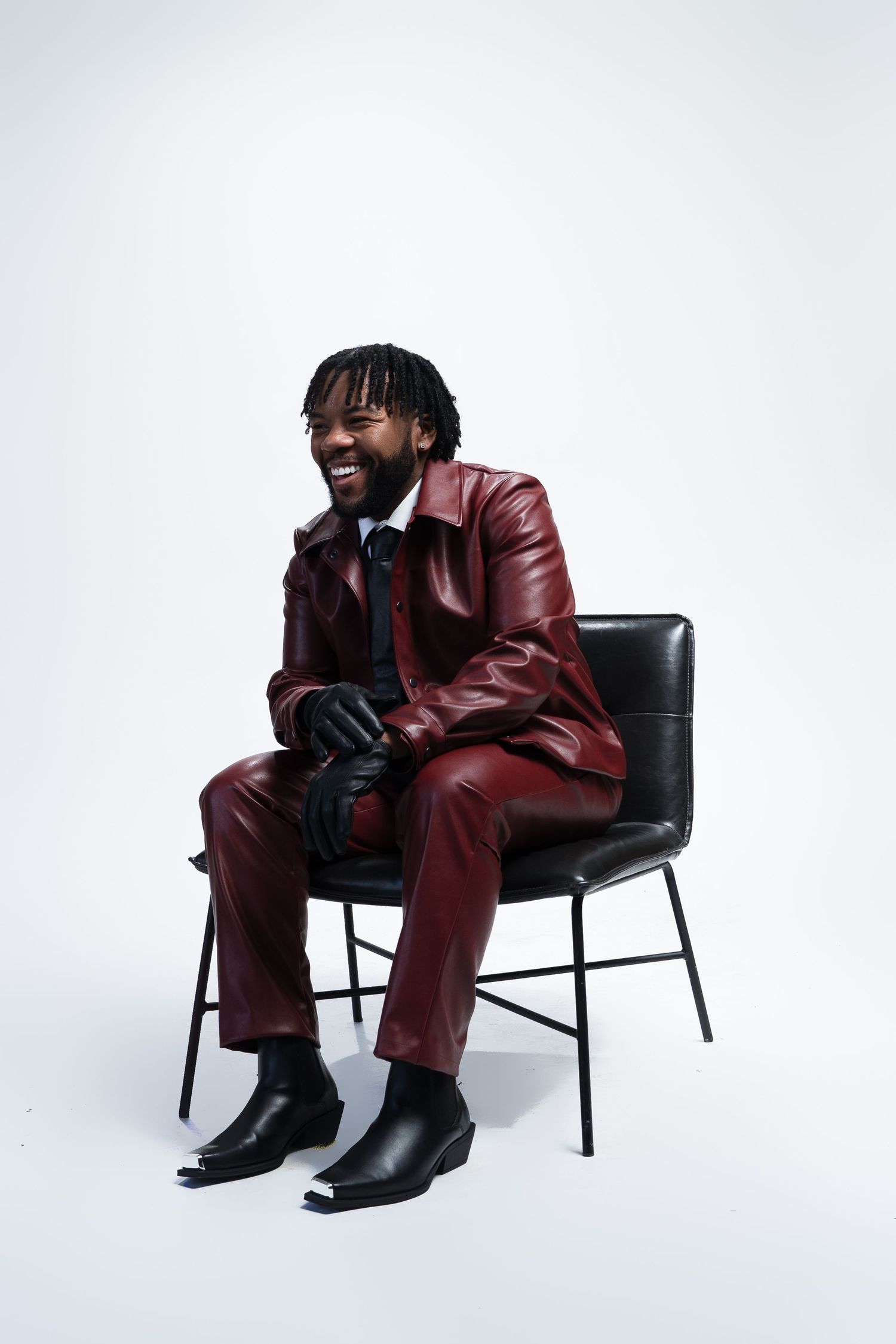 Figure in maroon leather suit laughing while seated on a black chair.