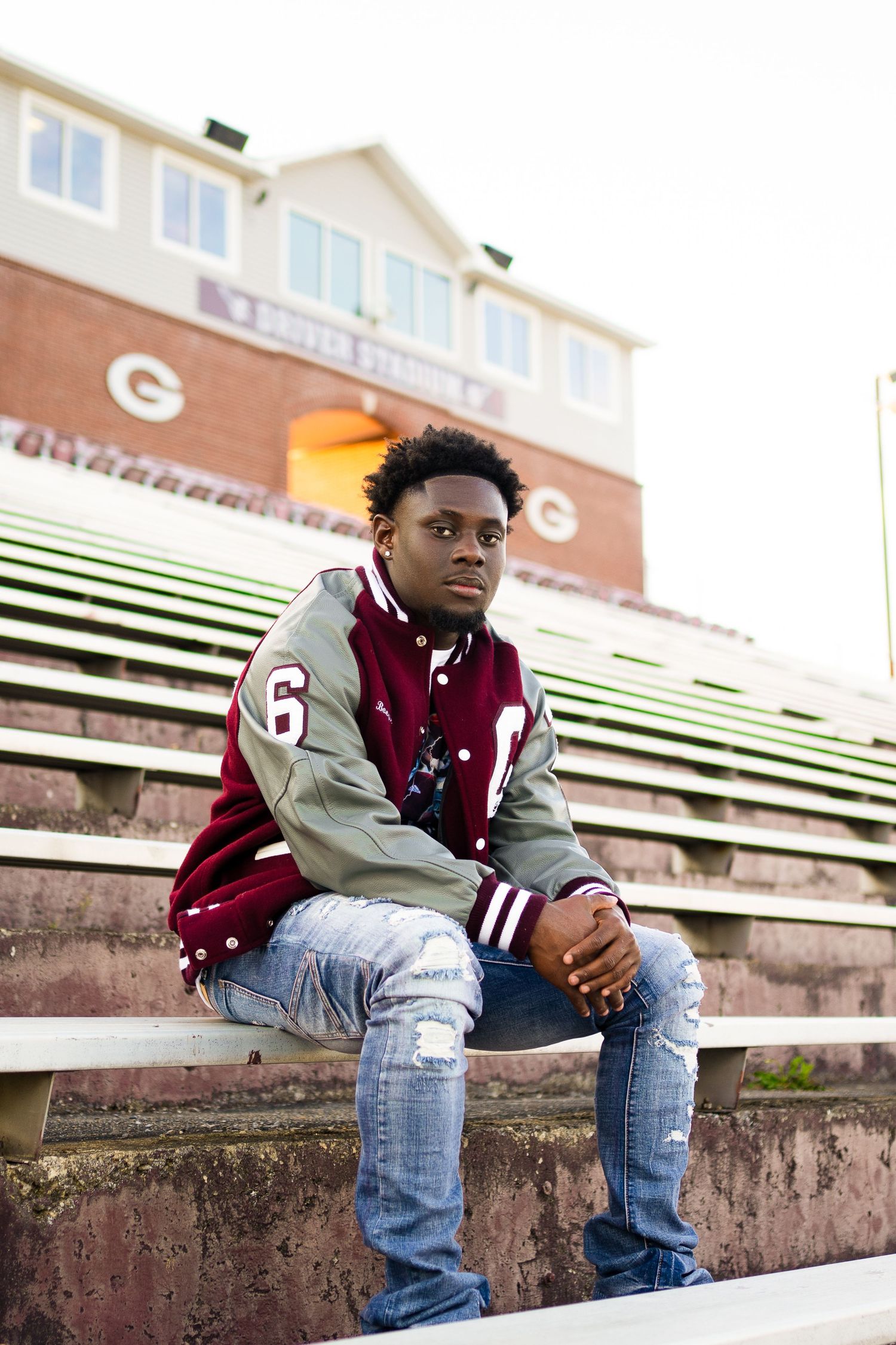 Athletic pose on stadium bleachers wearing varsity jacket and distressed jeans.