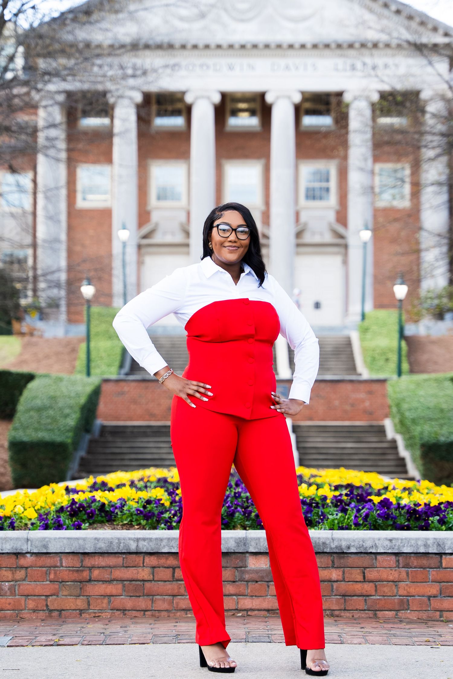 Vibrant red jumpsuit outfit photographed in front of a stately columned building with flower gardens.