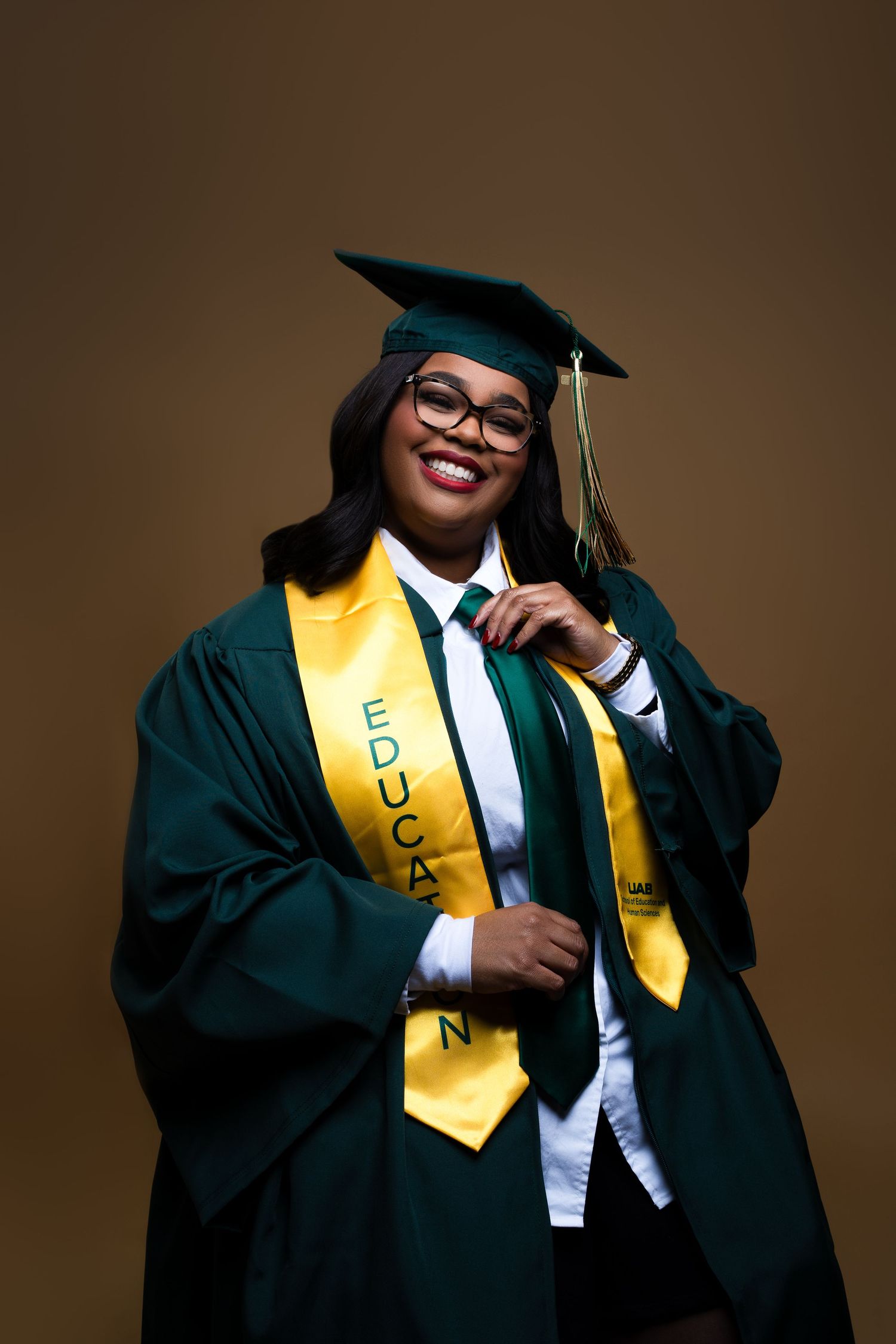 Graduate in green cap and gown with yellow education sash smiles while wearing glasses against brown background.
