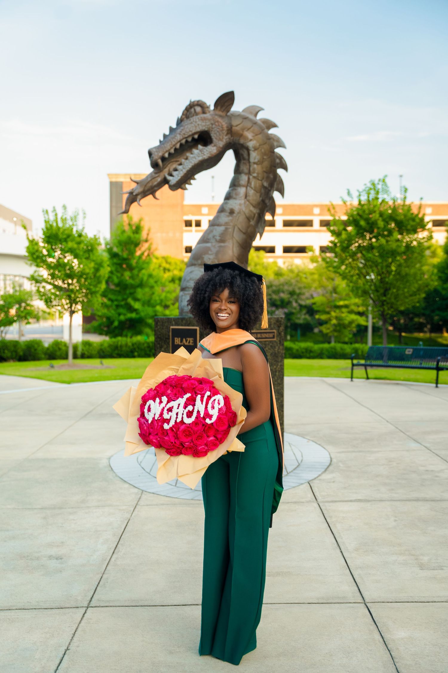 Graduate in green gown holds floral bouquet in front of dragon statue at university campus ceremony.