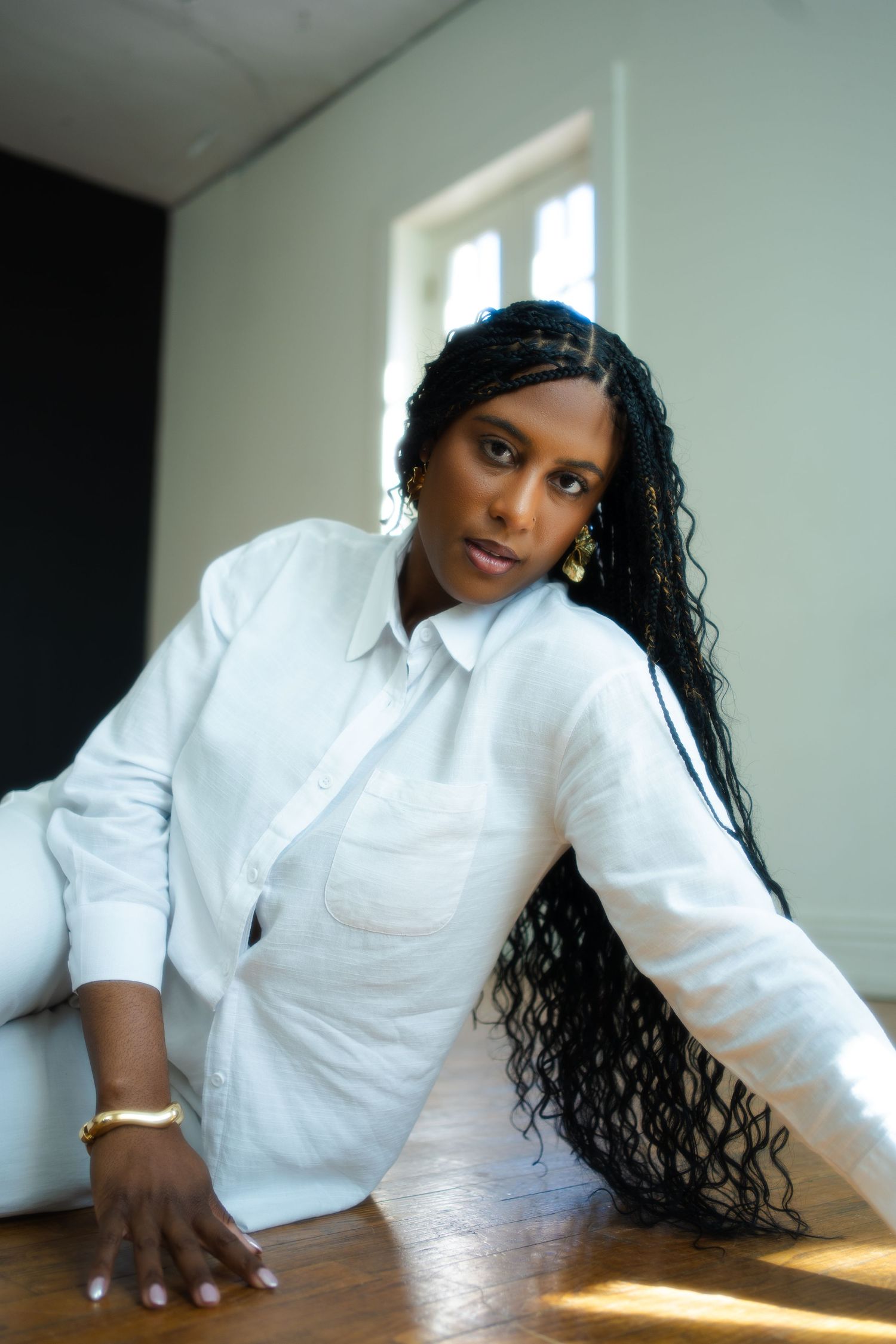 Woman in crisp white button-down shirt with long dark braids leans on wooden table in sunlit room.