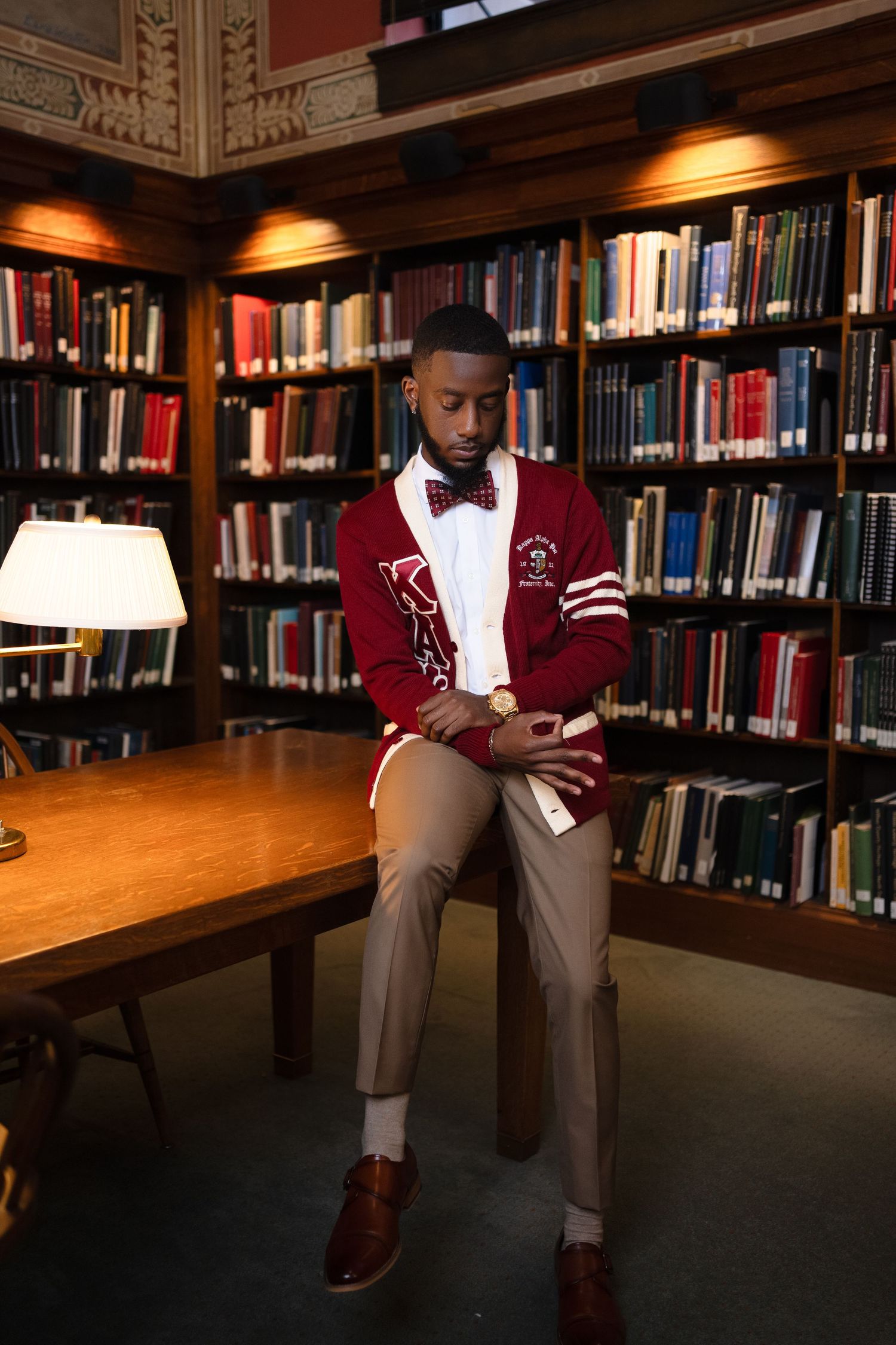 Graduate in varsity jacket sits thoughtfully in library among rows of books.