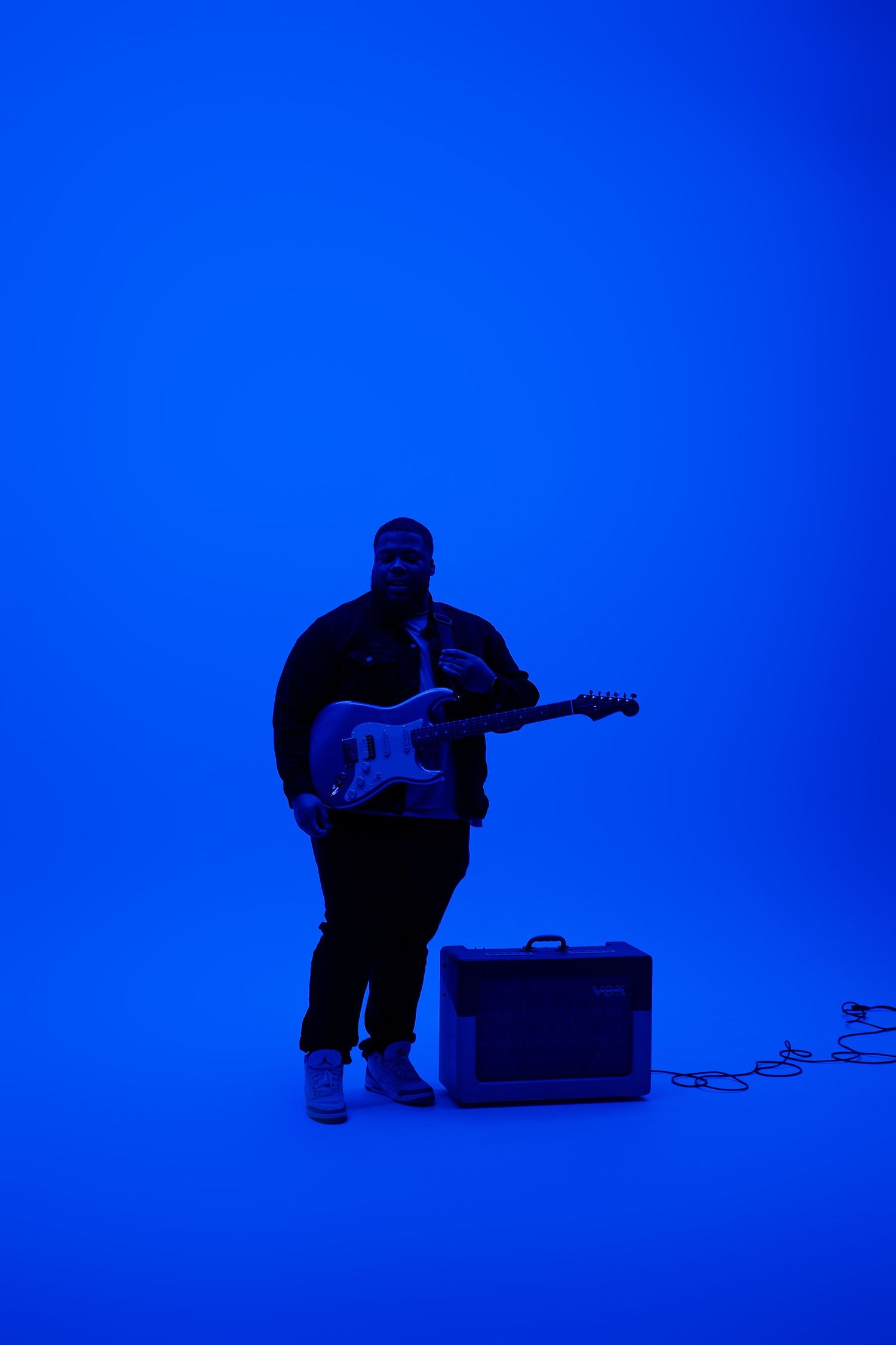 Musician with electric guitar and amp performing in blue-lit studio setting.