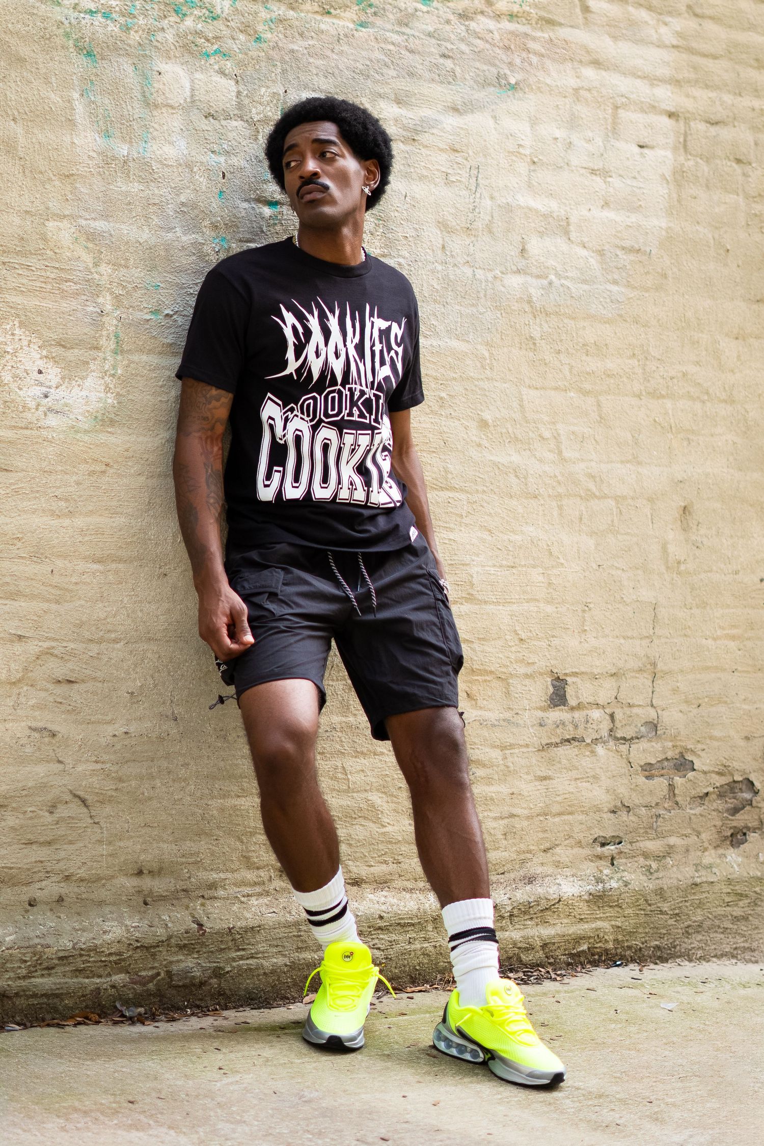Urban fashion photo showing person in black graphic tee, shorts and neon yellow sneakers against wall.