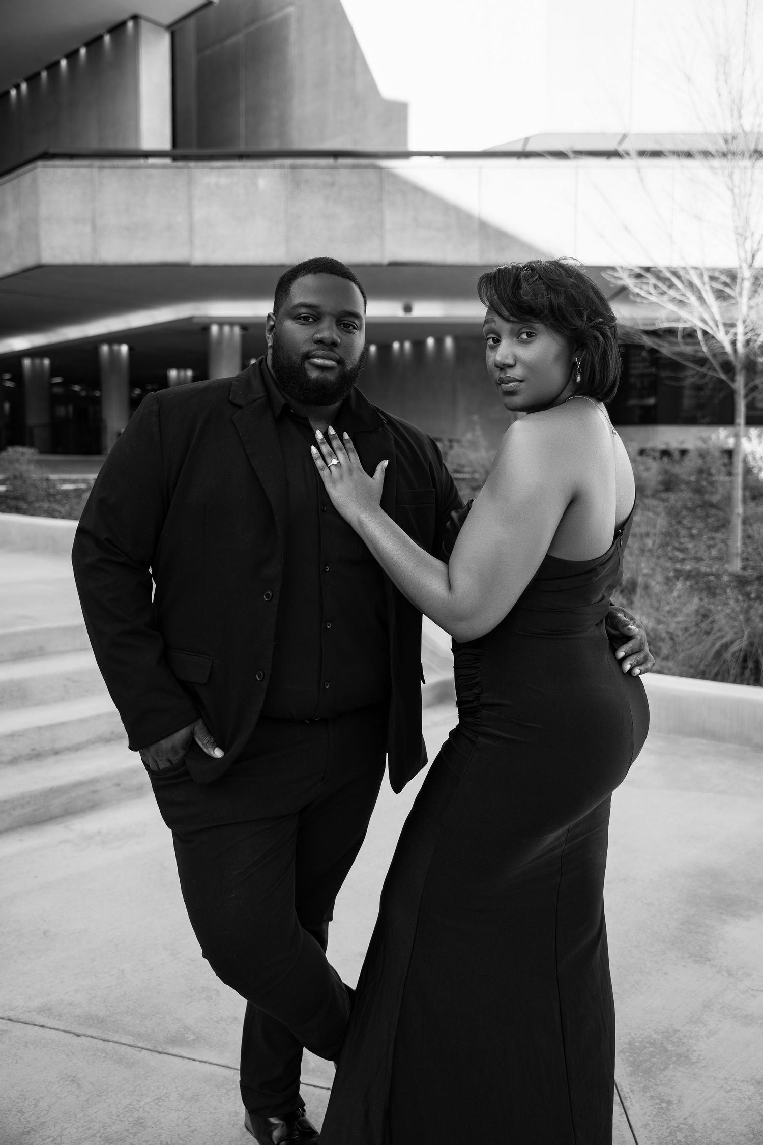 Black and white photo of an elegant couple in formal black attire posing outdoors near a modern building.