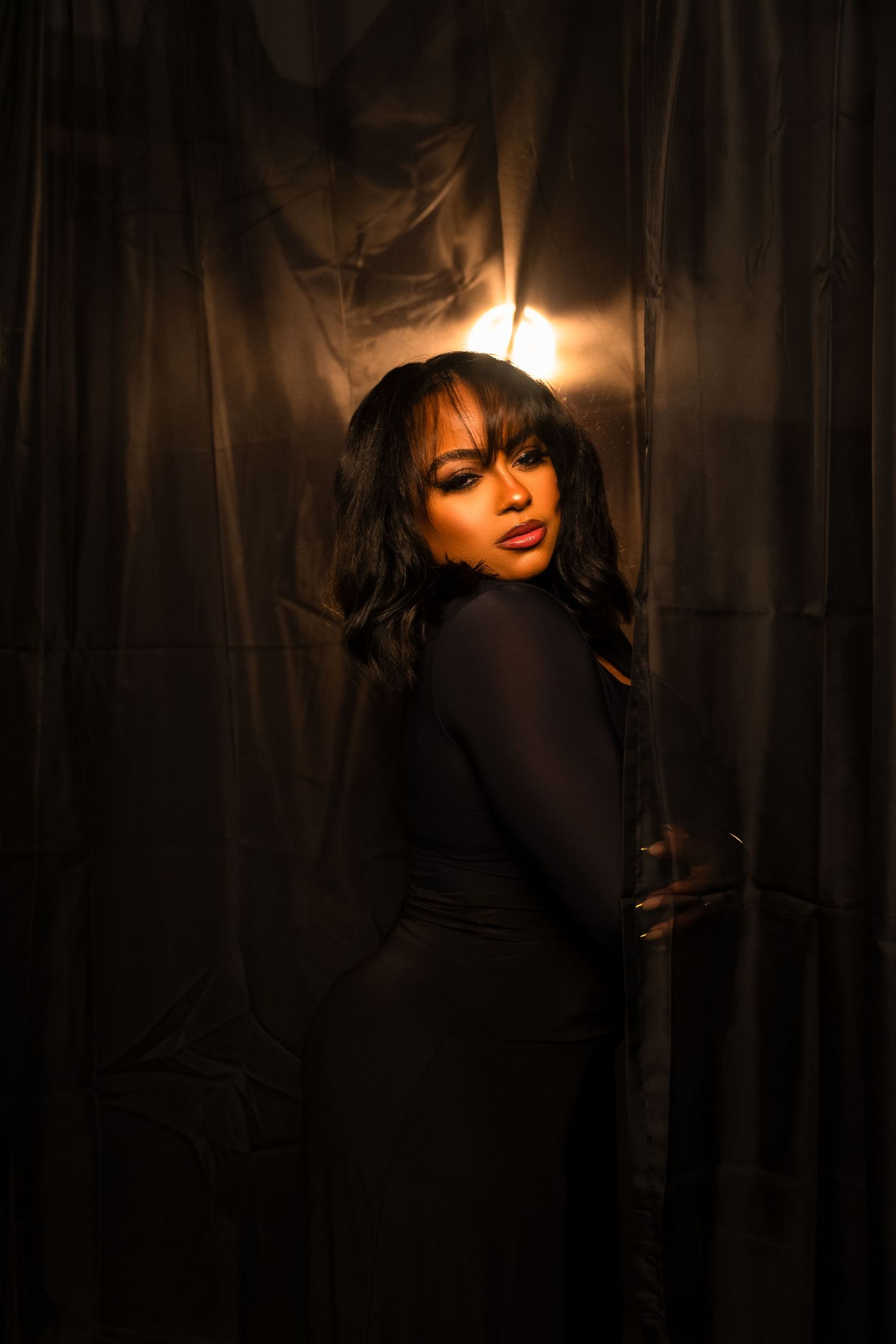 A woman in a black dress poses dramatically against dark metallic curtains with warm backlight.