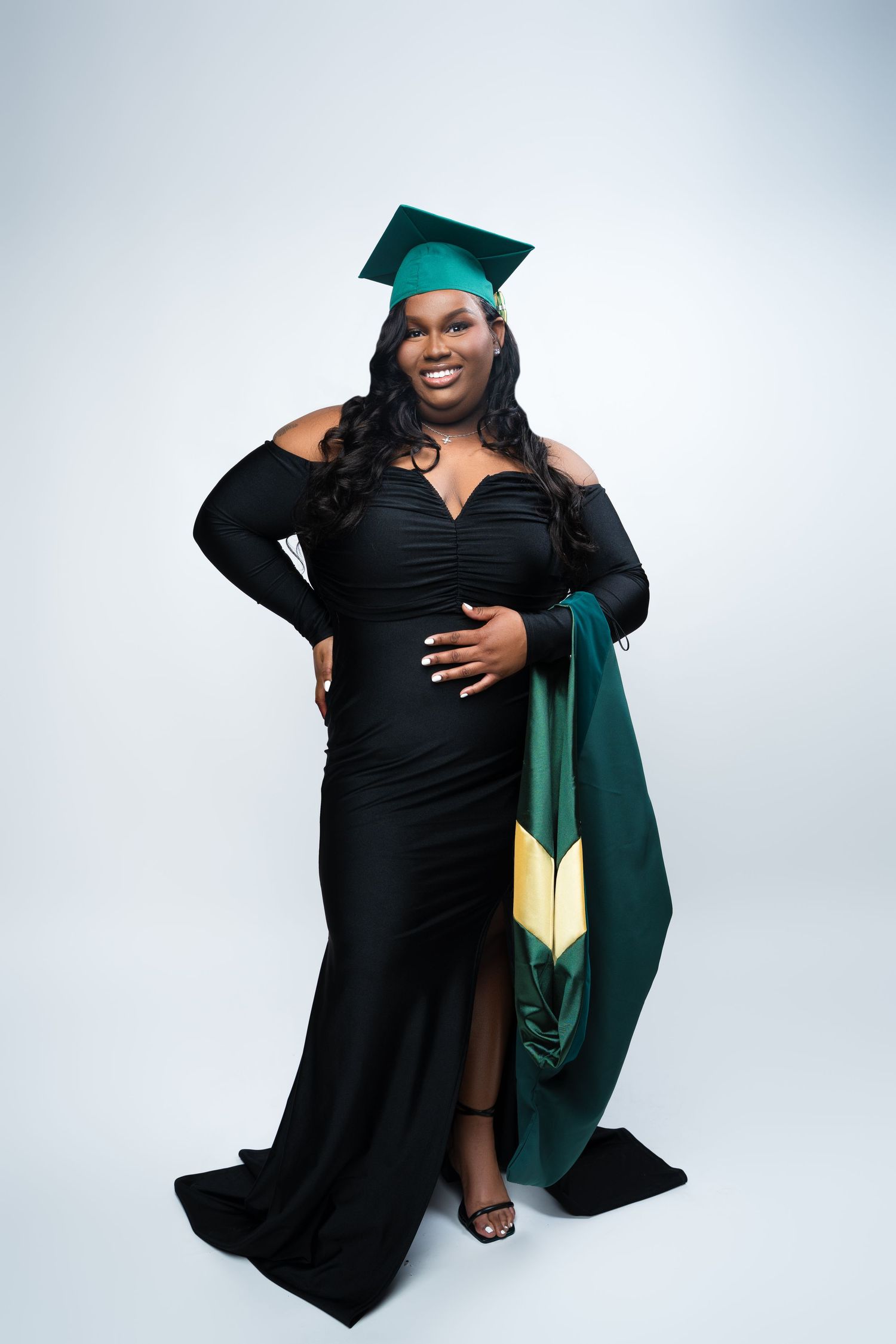 Pregnant woman in black gown and green graduation cap smiling, holding baby bump, wearing academic stole.