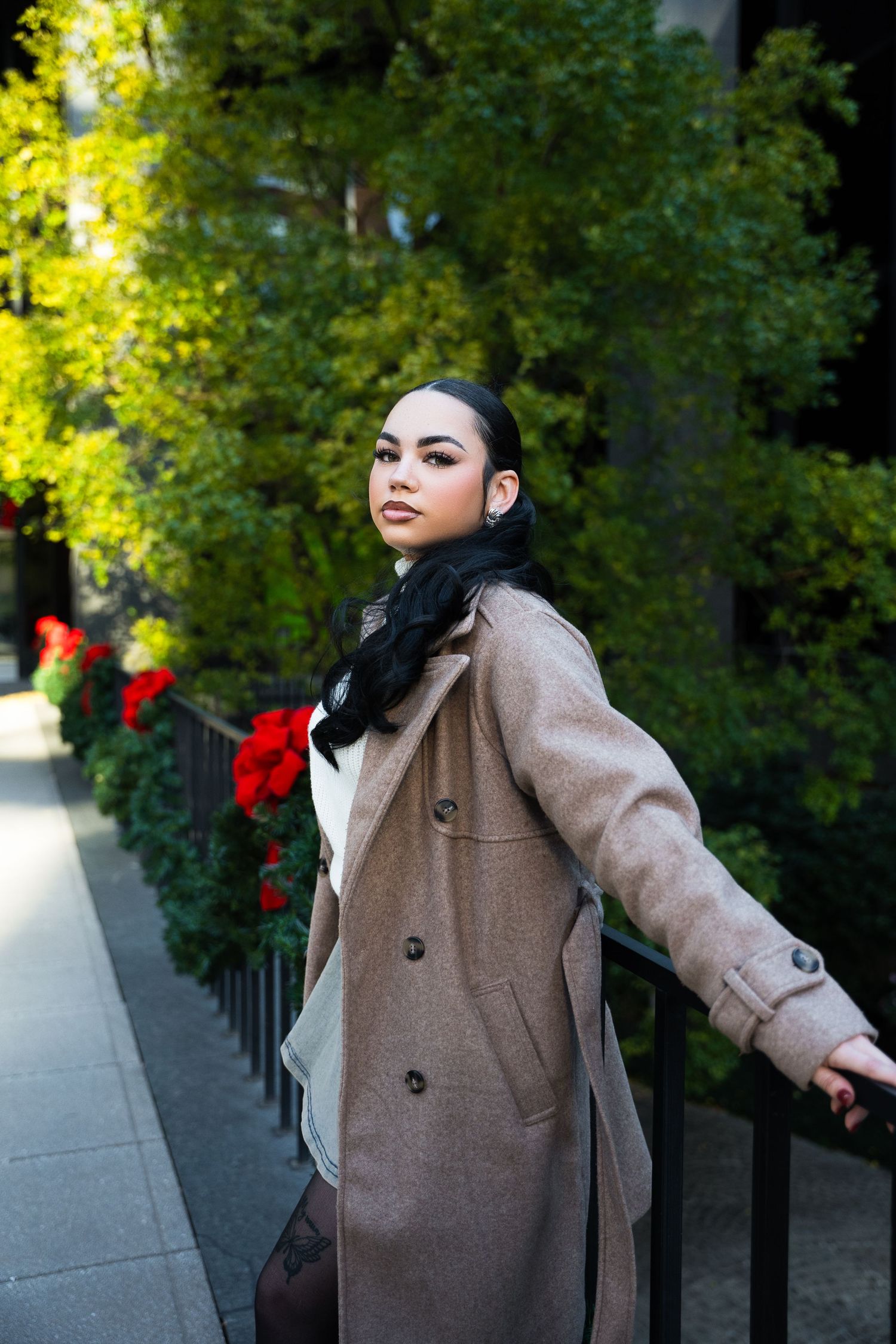 A person wearing a beige wool coat poses outdoors against a backdrop of green foliage and red flowers.