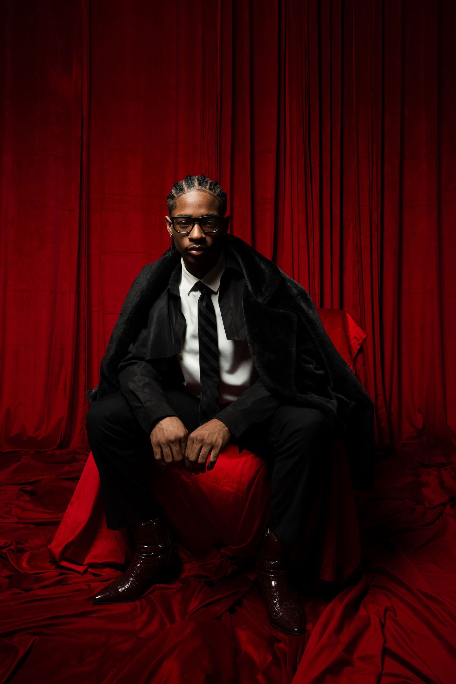 Figure in black suit sits on red fabric against dramatic red curtain backdrop in artistic portrait series.