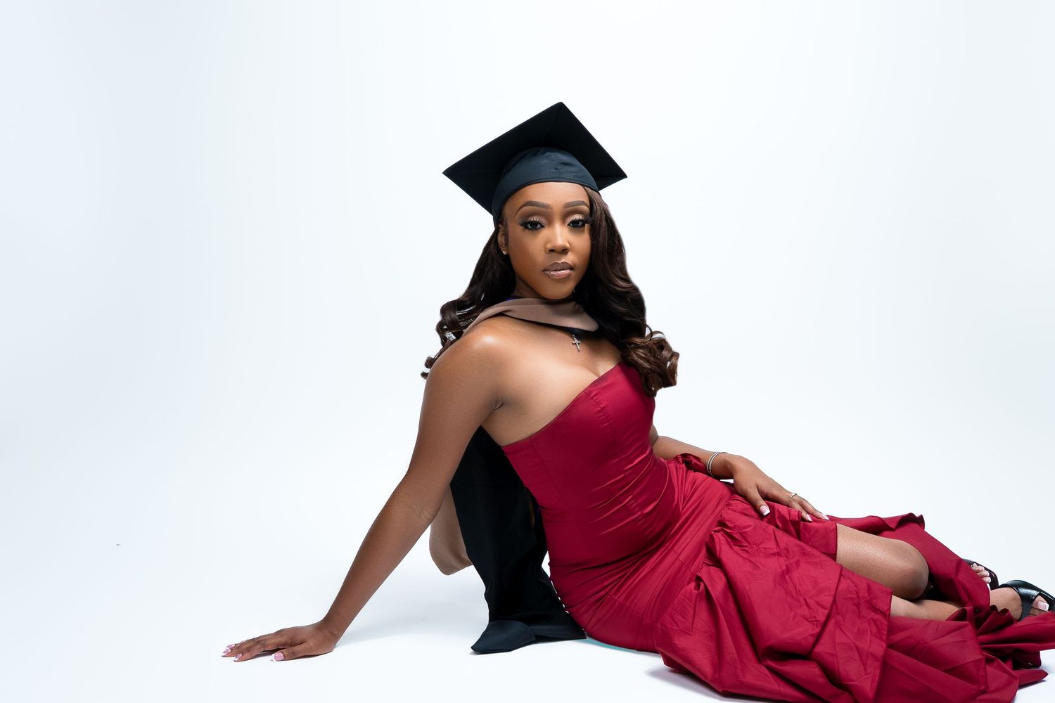 Graduate poses in red evening gown and graduation cap against white backdrop.