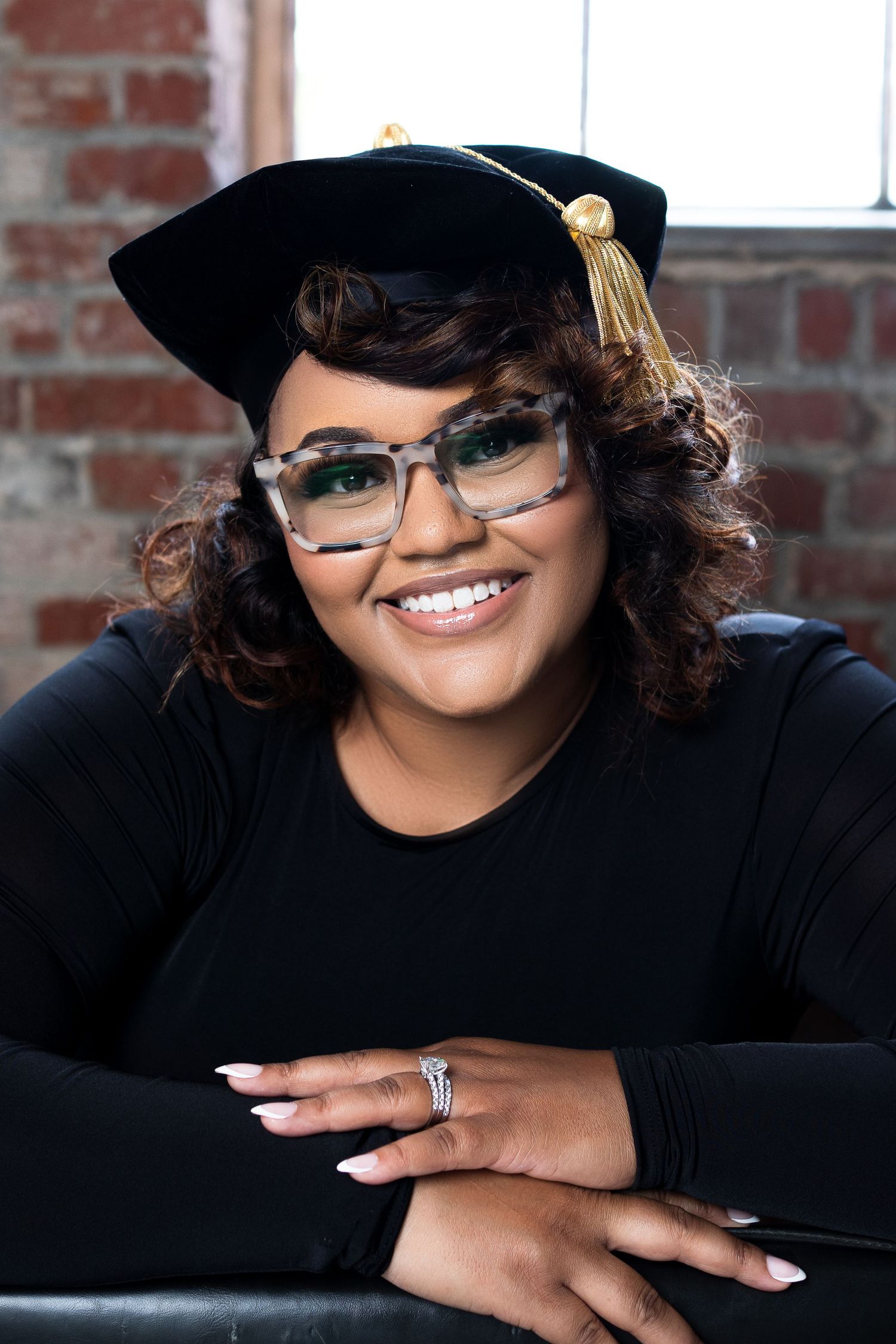 Professional headshot featuring stylish glasses, graduation cap and warm smile against brick wall background.