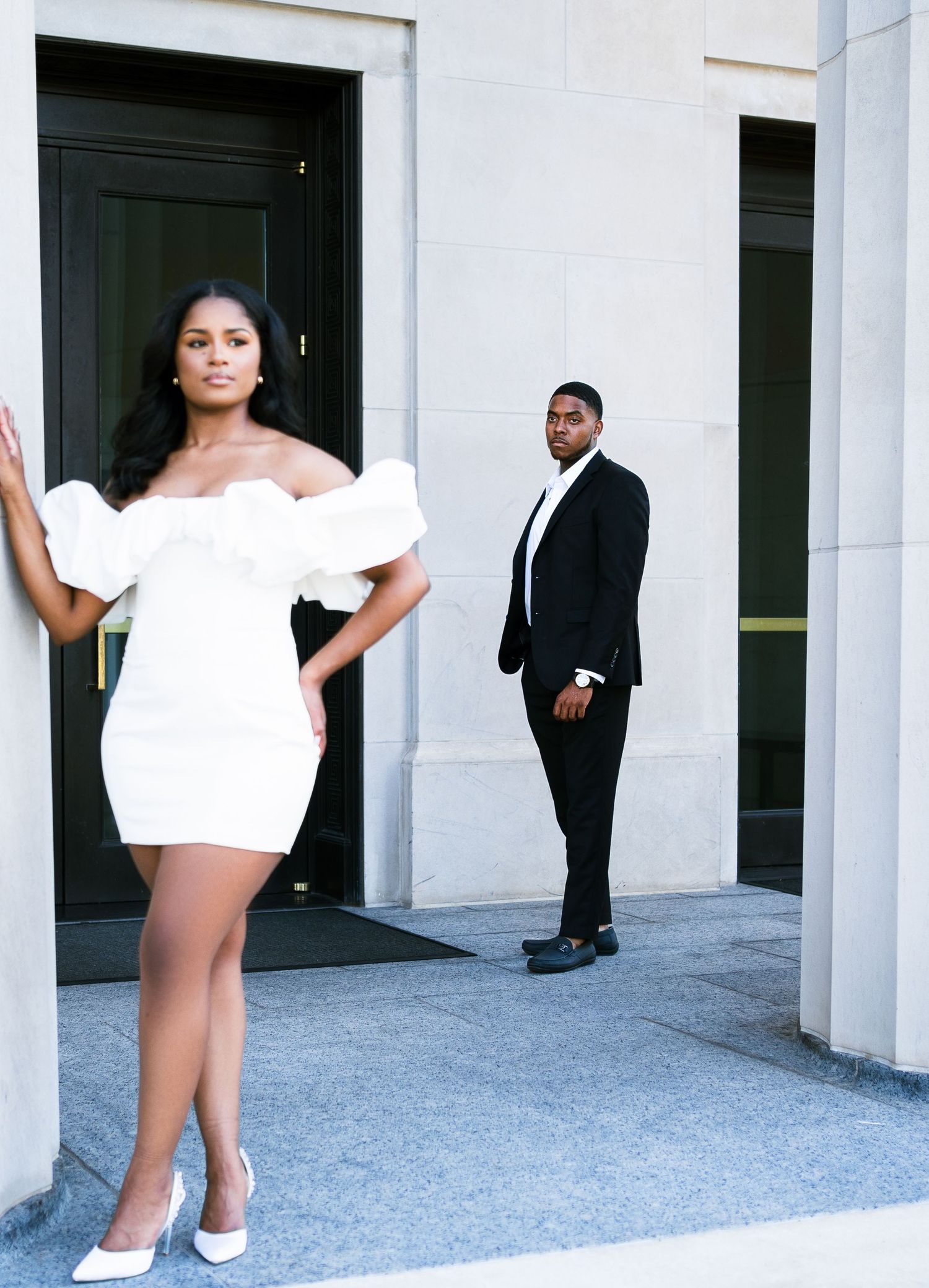 A couple poses separately in formal attire against a white building exterior.