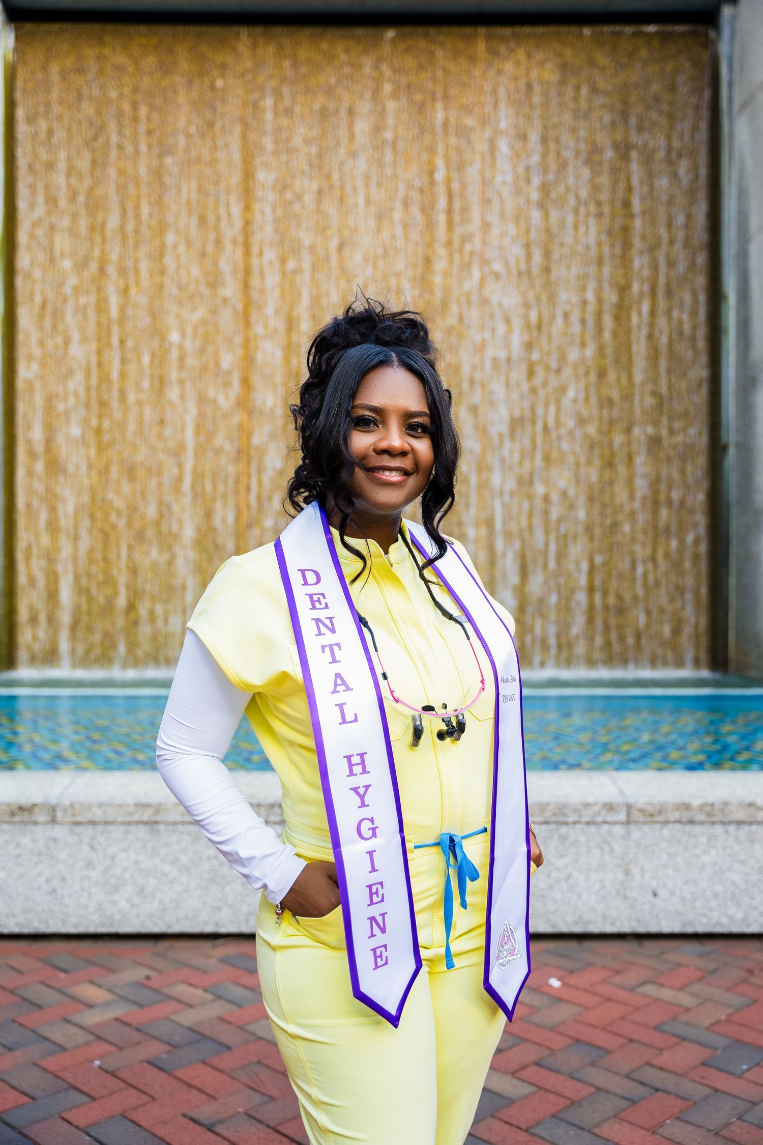 Graduate wearing yellow outfit and sash poses for graduation photo on campus.