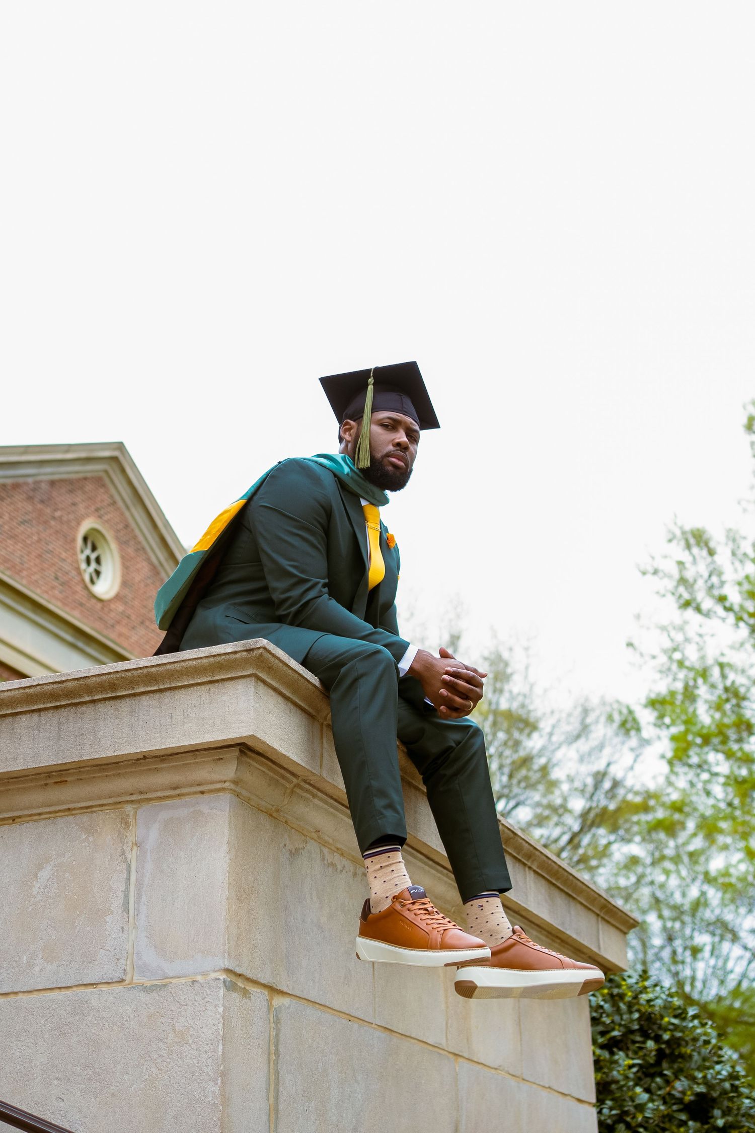 Graduate in green regalia sits on stone architecture during graduation photoshoot.