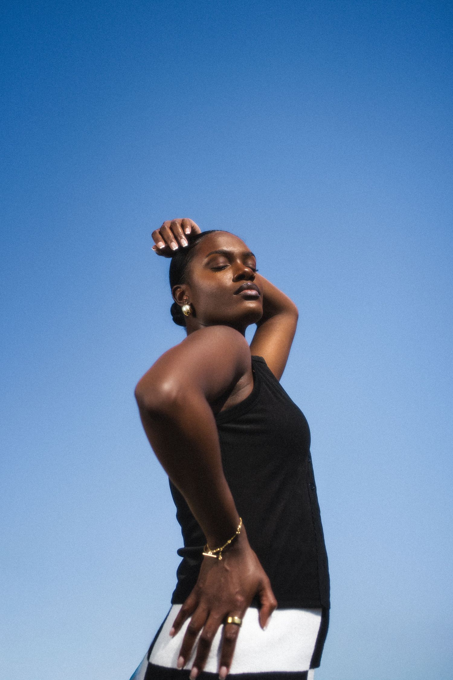 A person in a black tank top poses expressively against a bright blue sky background.